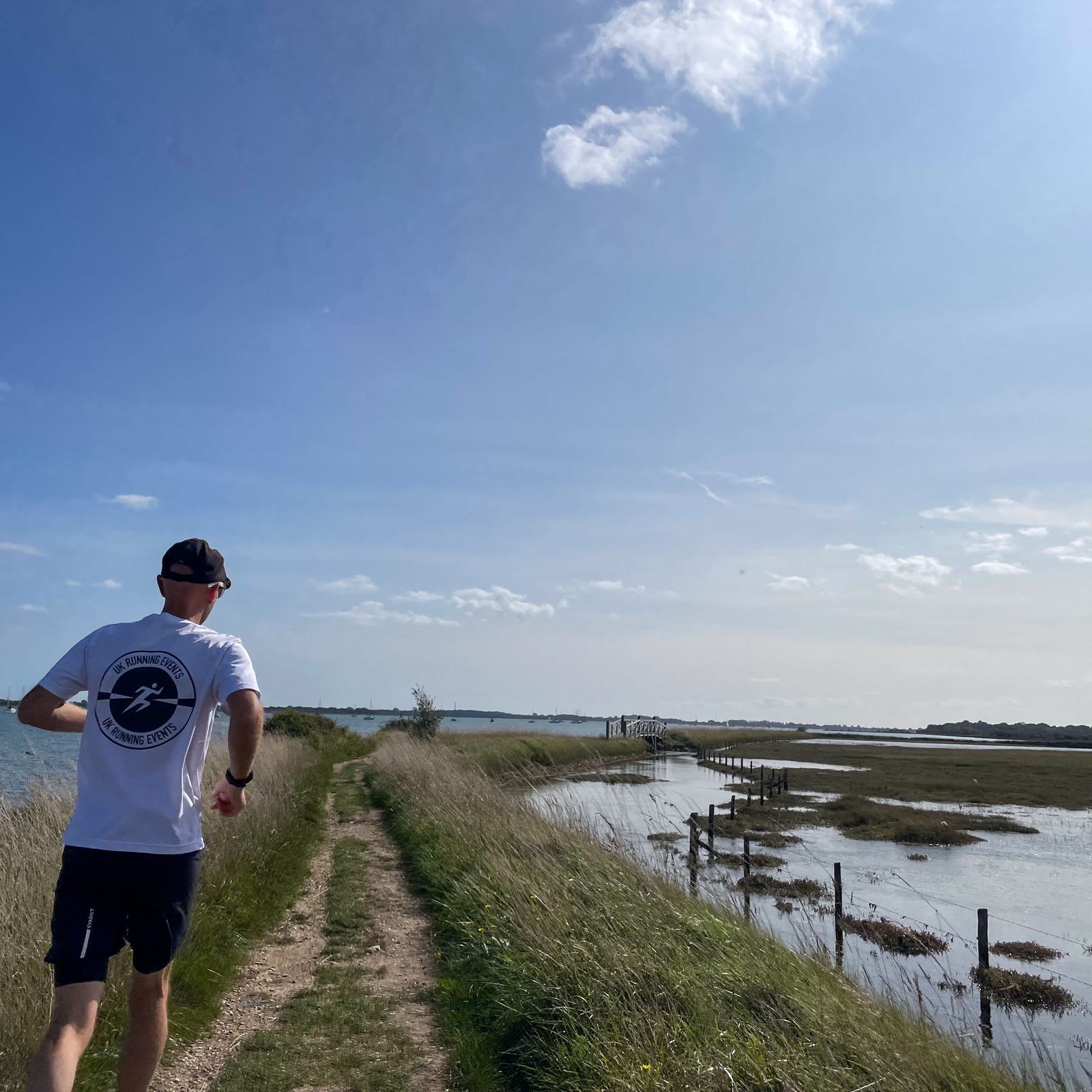 A person wearing a white shirt and black cap is jogging along a grassy path next to a body of water and marshland under a blue sky with scattered clouds.