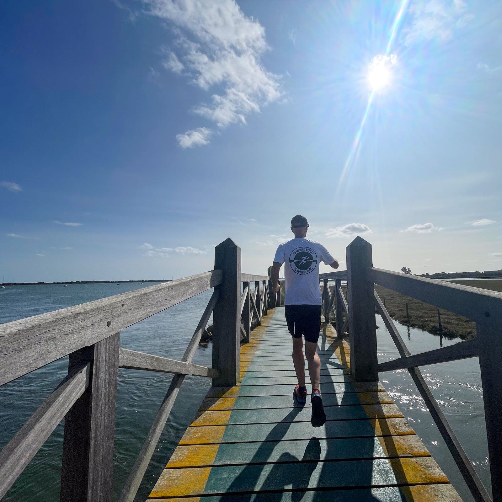 Person running on a wooden bridge over water, wearing a white shirt and black shorts. The sun is shining brightly in a blue sky with a few clouds. Shadows of the person and railings are cast on the bridge.