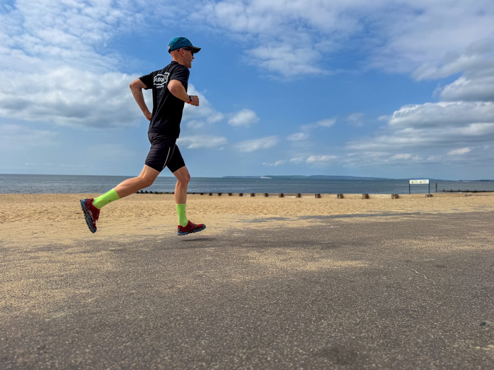 A person in athletic clothing and a teal cap runs along a paved path by the beach, with sand, sea, and a cloudy blue sky in the background.