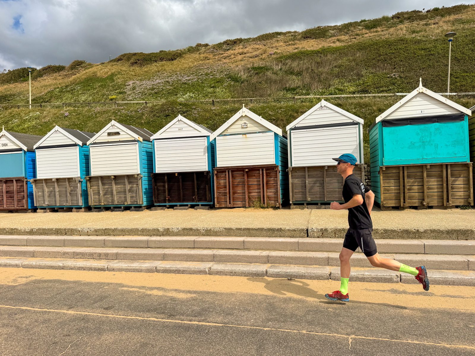 A man in athletic clothes jogs along a path in front of a row of colorful beach huts with green grassy hills and a cloudy sky in the background.