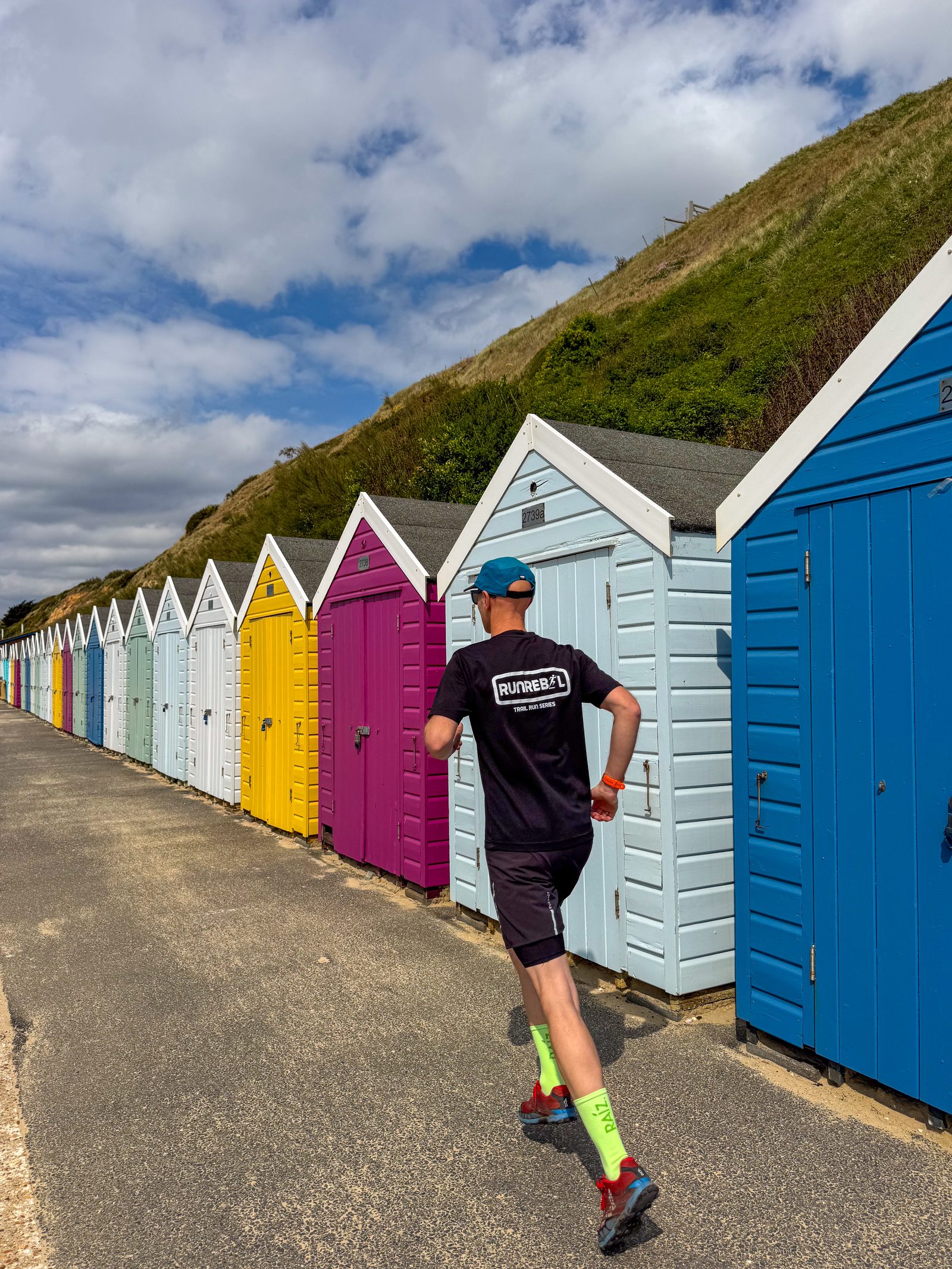A person in running gear jogs along a row of colorful beach huts on a sunny day, with a grassy hillside and blue sky with scattered clouds in the background.
