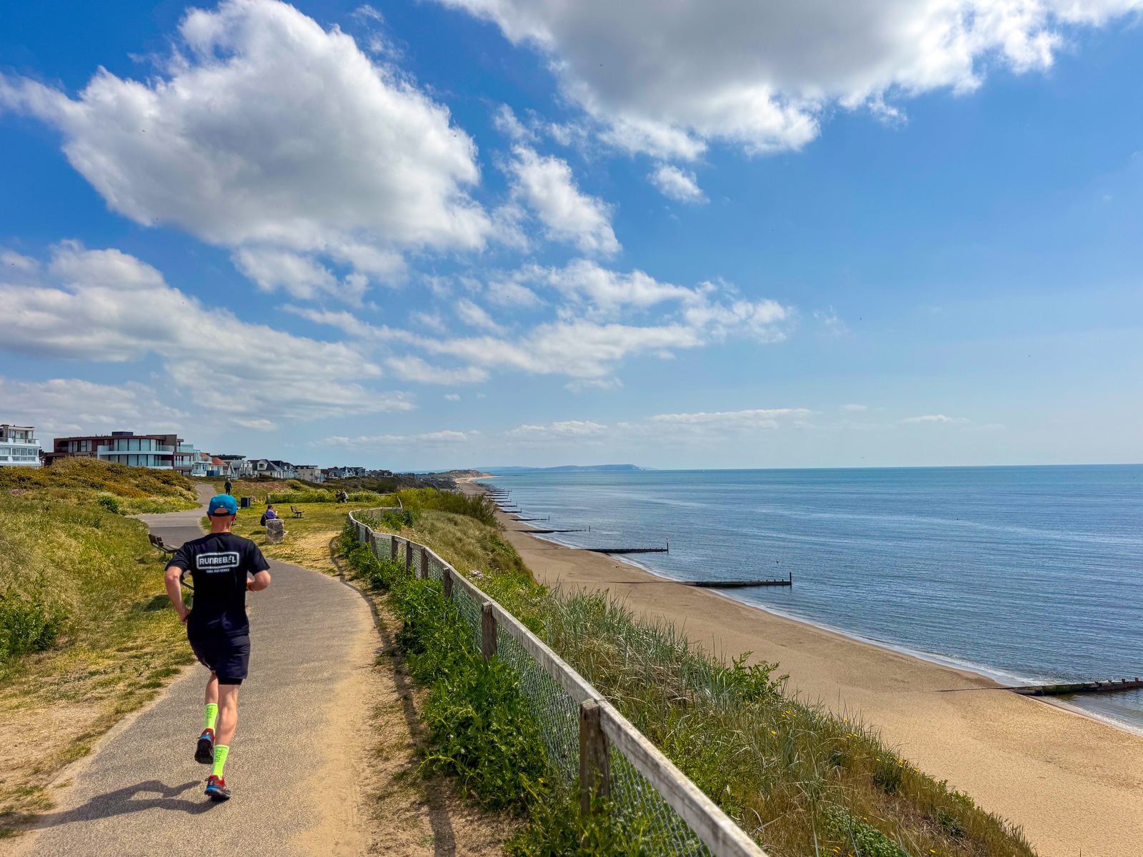 A person in athletic clothing runs on a paved coastal path beside a sandy beach, with calm sea and houses in the distance under a partly cloudy blue sky.