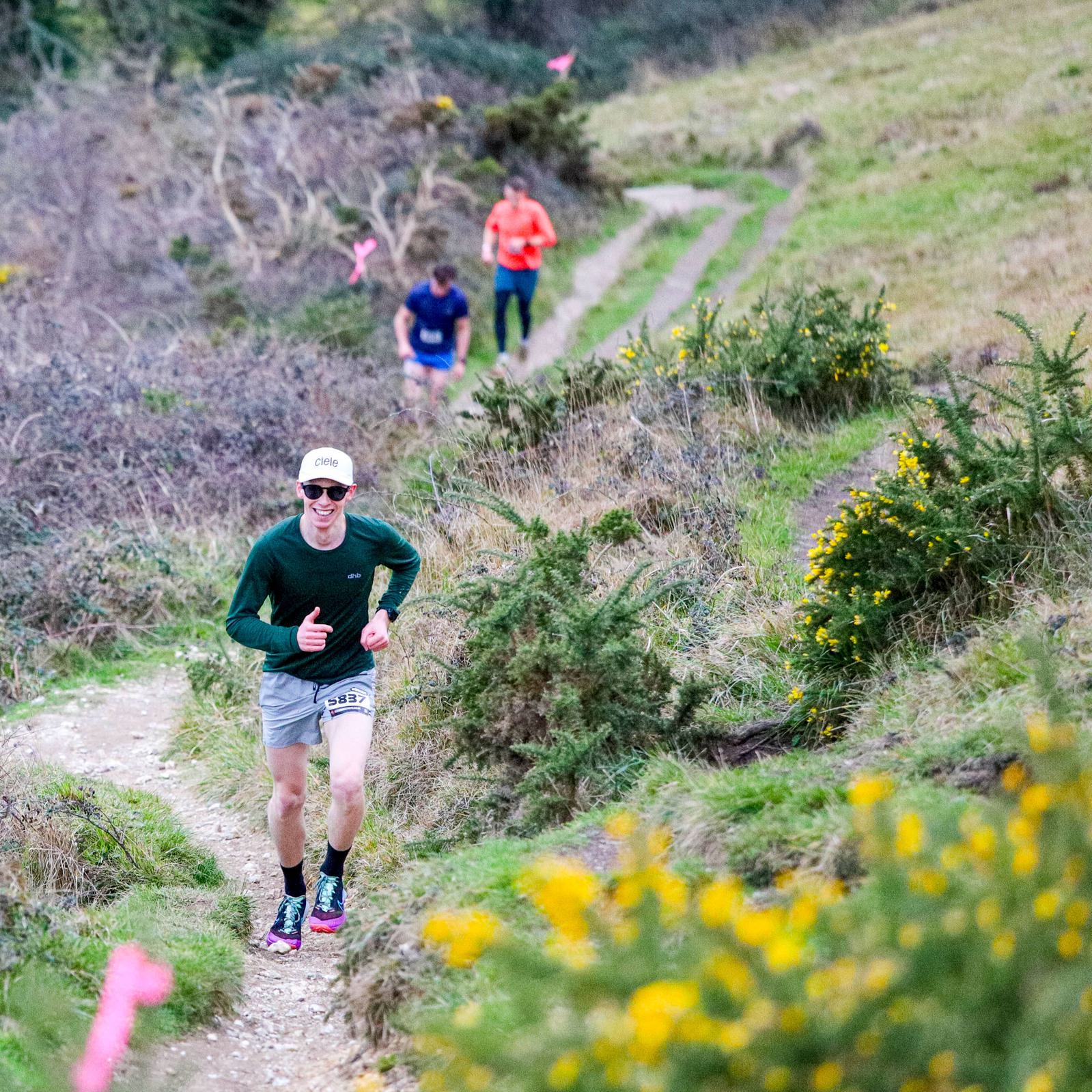 A person in a green long-sleeve shirt and cap runs on a grassy trail surrounded by bushes and yellow flowers. Two more people follow on the winding path. The scenery includes a hilly landscape with some trees in the background.