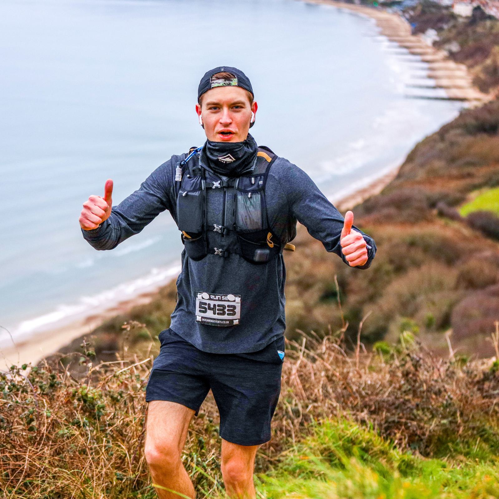 A man running outdoors gives two thumbs up. He's wearing a cap, a hydration pack, and a race bib with the number 5433. He is running on a trail with a coastal landscape in the background, featuring cliffs and the sea.
