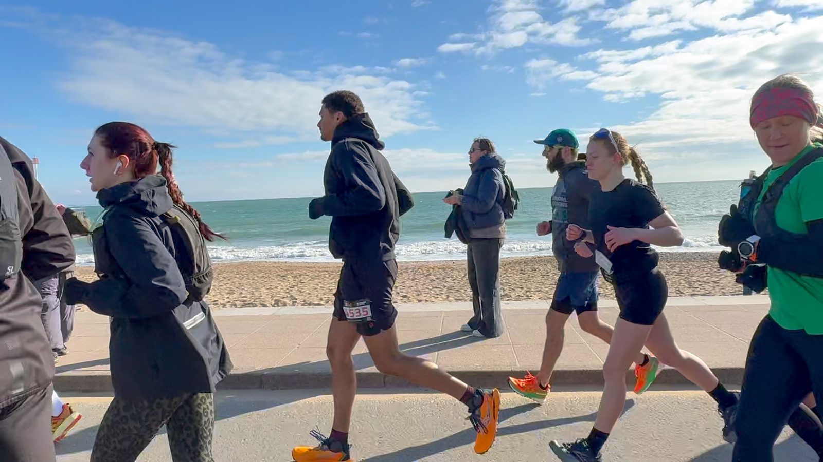 Runners jog along a seaside promenade on a sunny day, with the beach and ocean in the background. A few people stand watching, dressed warmly. The sky is partly cloudy.