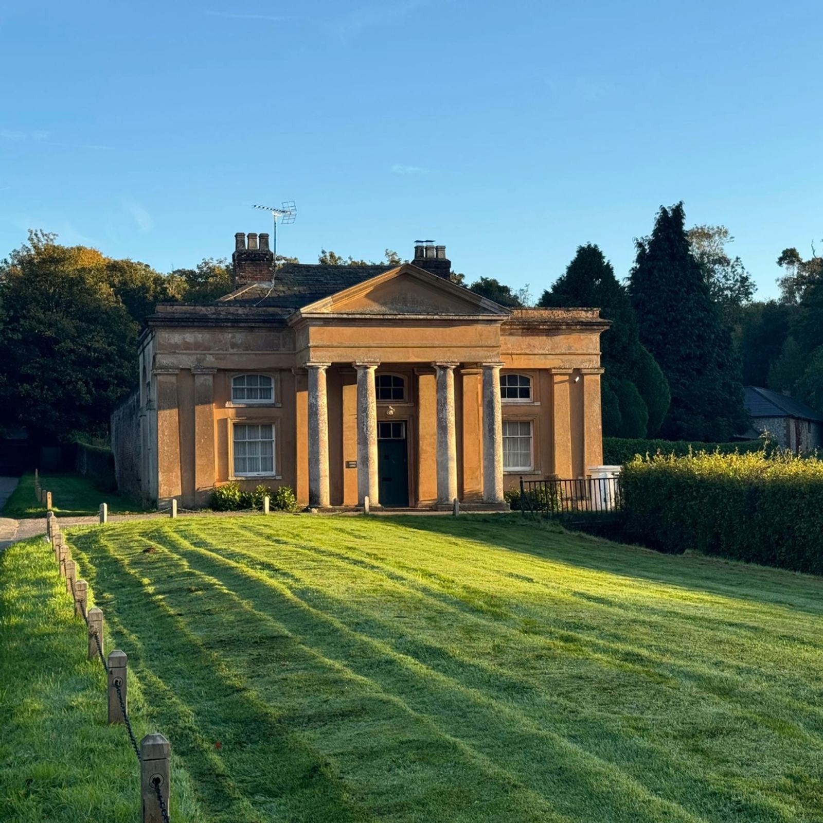 A small, classical-style building with four columns in front stands in the sunlight. It's surrounded by lush greenery with a neatly mowed lawn in front, a dirt path to the left, and trees in the background under a clear blue sky.