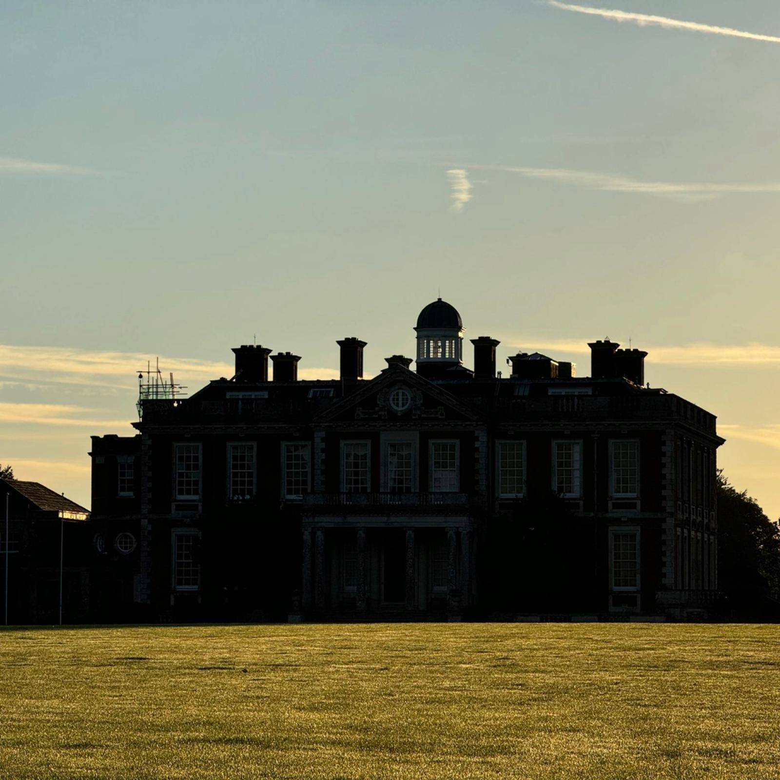 Silhouette of a large historic building with a dome and chimneys against a golden sky. The foreground is a sunlit, grassy field. Wispy clouds streak across the sky.