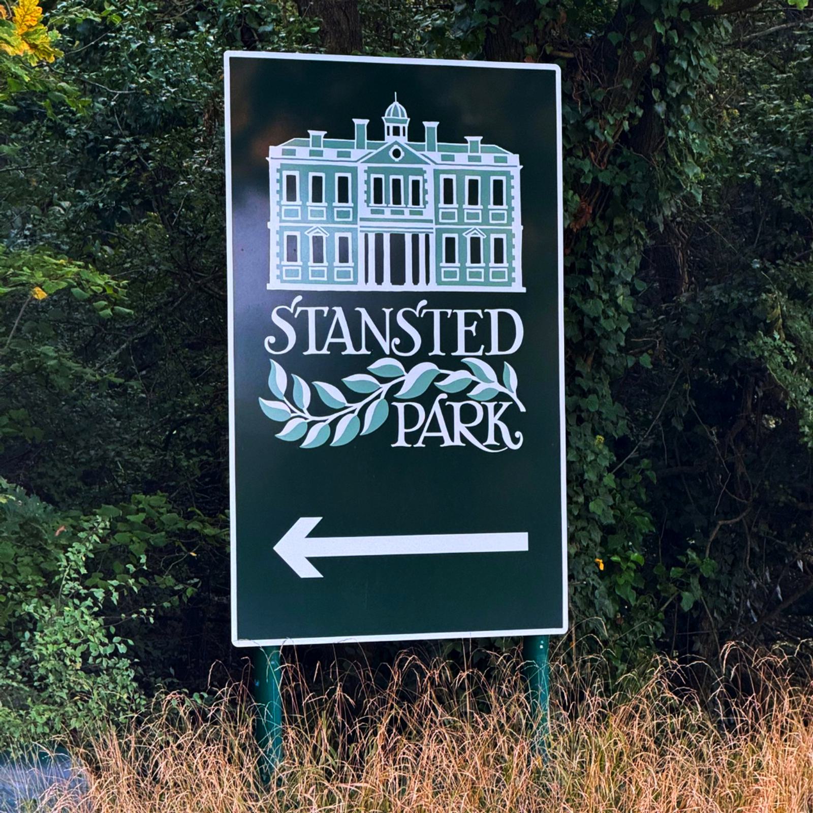 A green sign features a white illustration of a large building above the text "Stansted Park" with decorative leaves. An arrow points left. The sign is surrounded by tall grass and lush greenery.