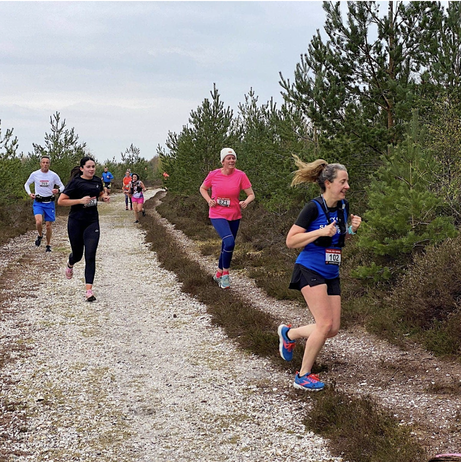 A group of people running on a gravel path through a forested area. The runners are wearing athletic gear and appear focused. The background features trees and cloudy skies, creating an outdoor race atmosphere.
