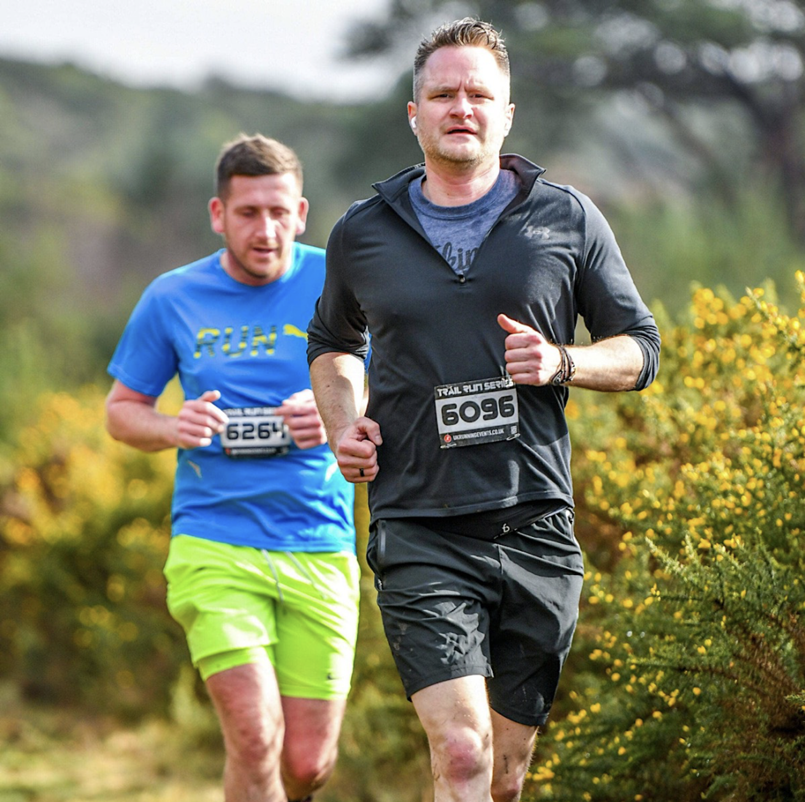 Two men running a trail race outdoors, surrounded by greenery. The man in the foreground wears a black outfit with the number 6096. The man behind him wears a blue shirt and yellow shorts with the number 6264. They appear focused and determined.