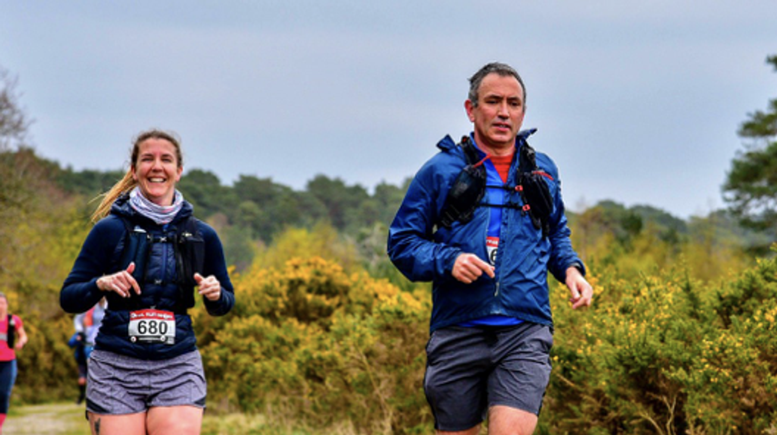 Two people are running outdoors on a trail, surrounded by greenery. The woman on the left wears a dark jacket and has a race bib numbered 680, smiling as she runs. The man on the right wears a blue jacket, appearing focused. Other runners are in the background.
