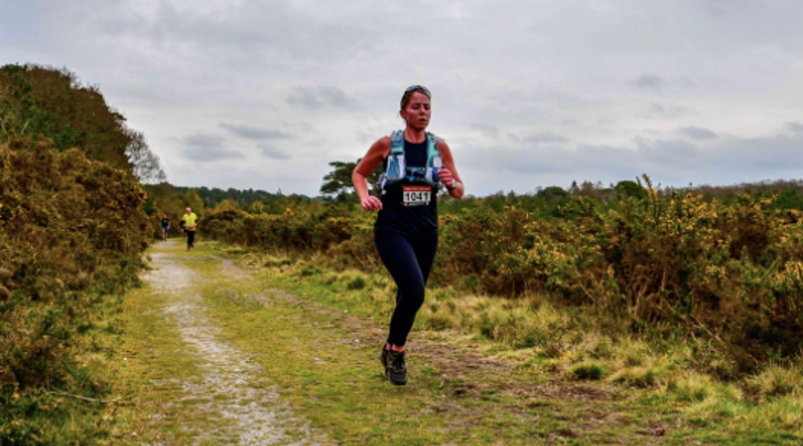A person in athletic gear is running on a dirt trail through a bushy landscape. The sky is overcast, and another runner is visible in the distance.