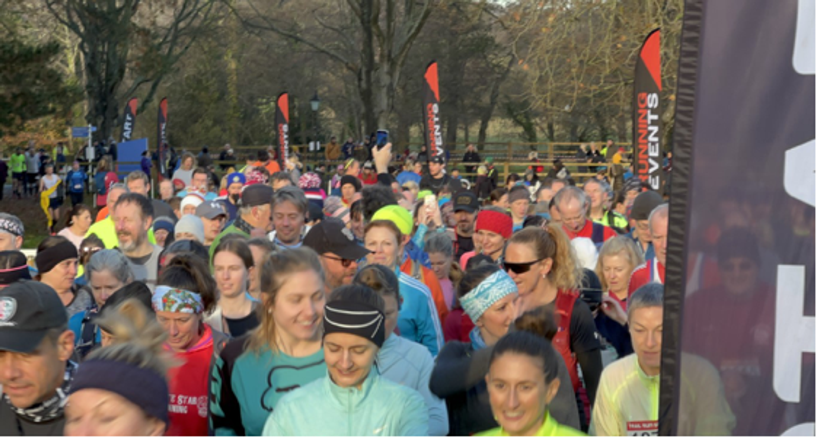 A large group of people participating in an outdoor running event, surrounded by trees. Many are wearing colorful athletic gear. Tall event flags are visible in the background. The mood appears lively and energetic.