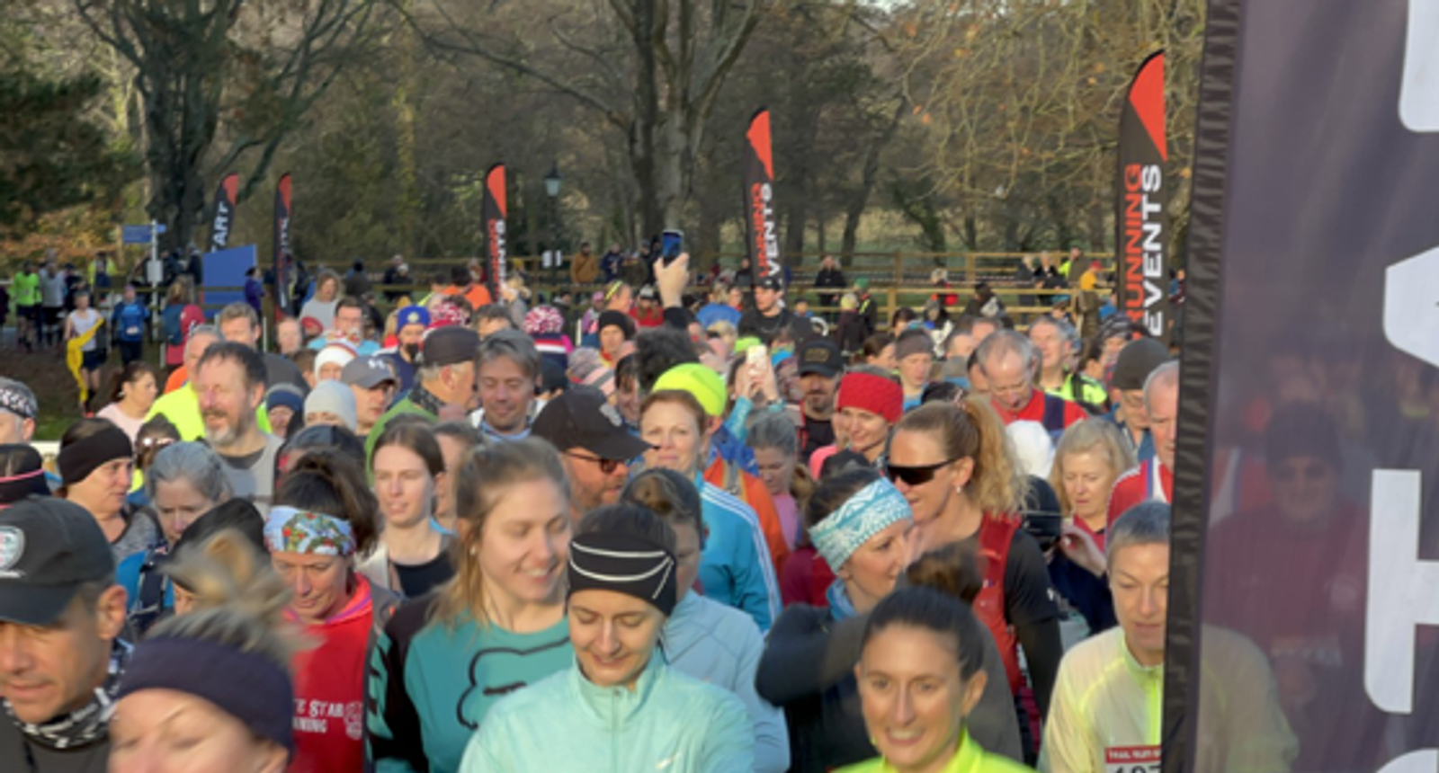 A large group of people participates in an outdoor running event, wearing various athletic gear. Trees and event flags are visible in the background. The atmosphere is lively and energetic.