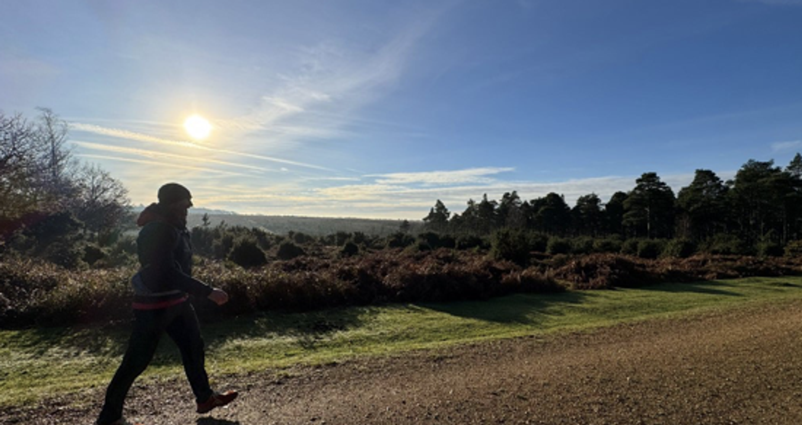 A person walks along a dirt path in a vast, open landscape with trees and bushes. The sun shines brightly in a clear blue sky, casting long shadows across the ground. The scene is peaceful and serene.