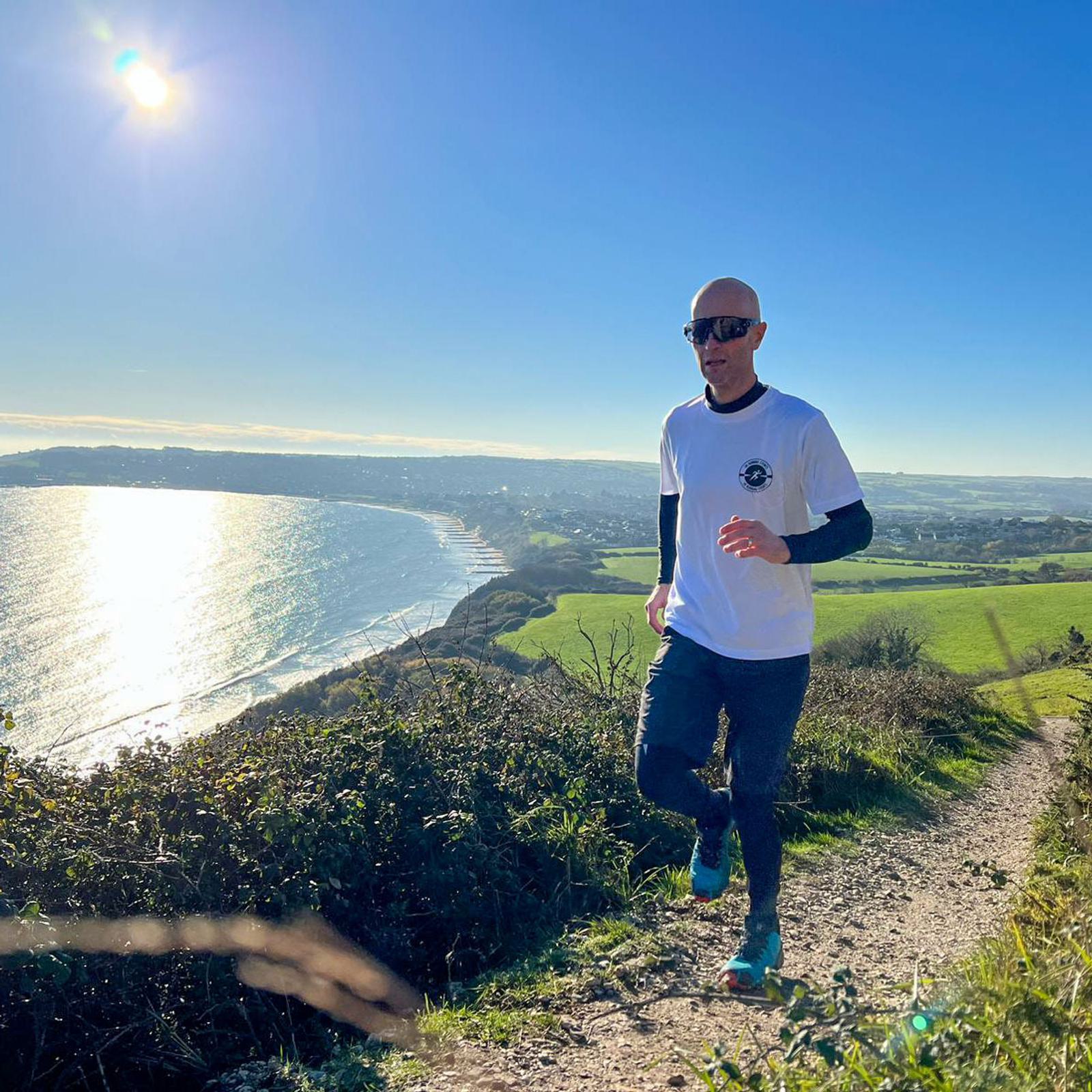A person in sunglasses and athletic clothing is running on a scenic trail. The path winds through grass and bushes with a view of the coastline and ocean under a clear blue sky and bright sun.