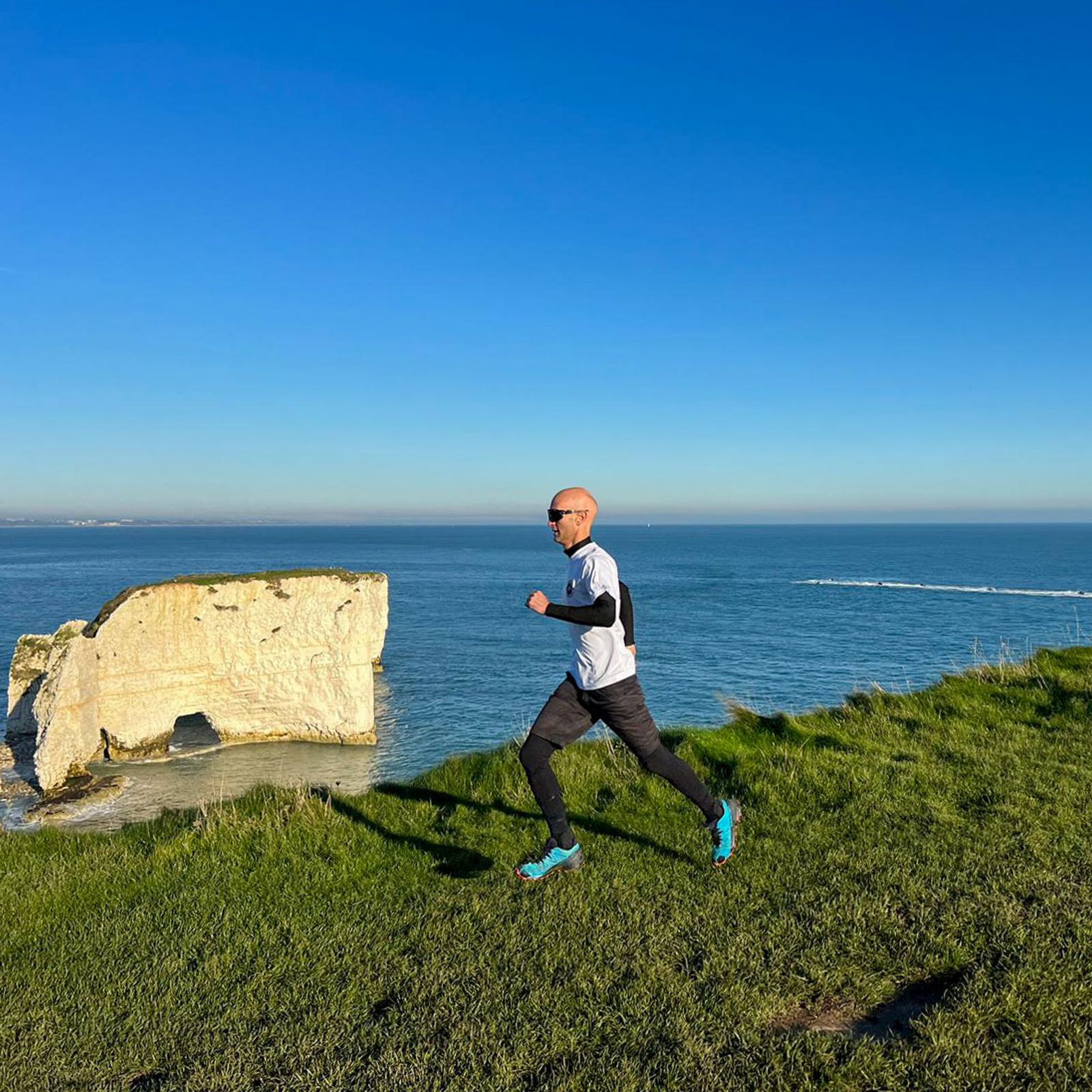 A person jogs on a grassy cliffside path overlooking the ocean. They are wearing a black and white athletic outfit, sunglasses, and blue running shoes. In the background, there is a large rock formation and a clear blue sky.
