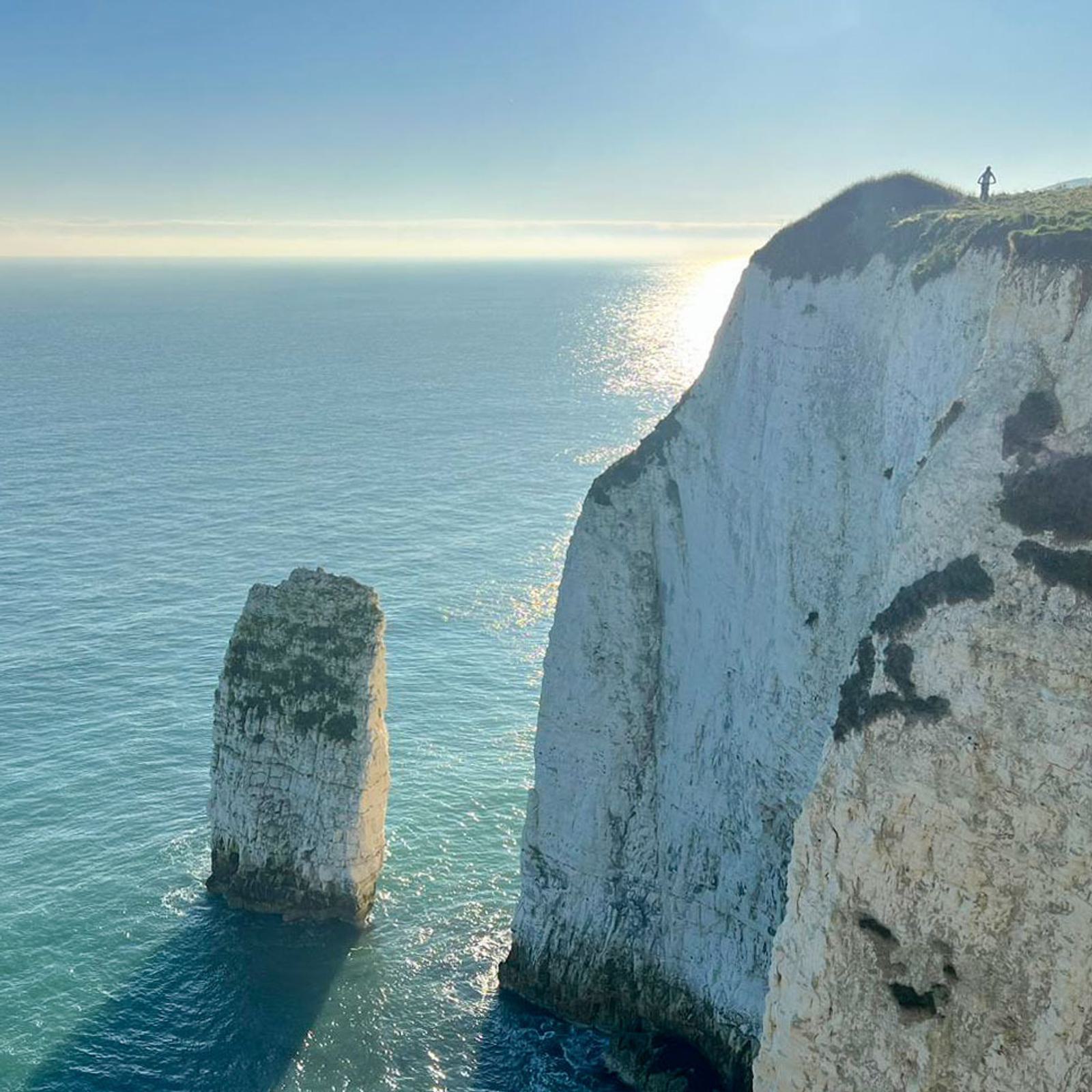 Clifftop view overlooking the ocean with a lone person standing on the edge. The scene features a towering white cliff and a solitary sea stack rising from the water. The sun casts a bright reflection on the calm sea.