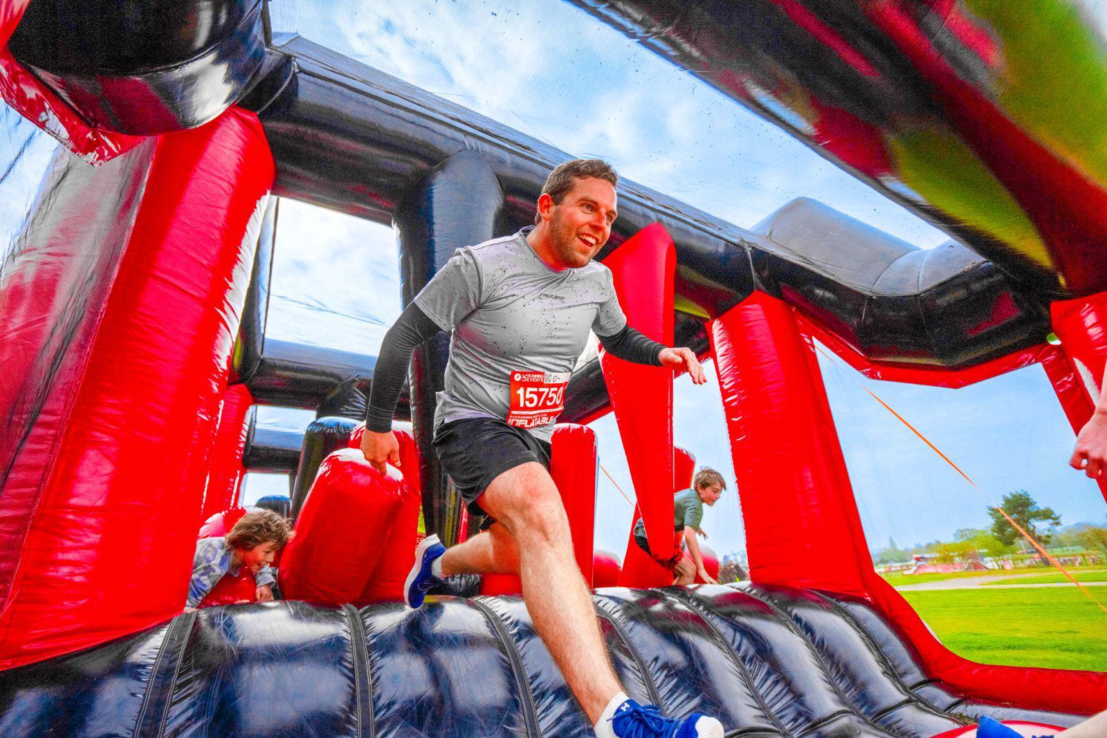A man in a gray shirt and black shorts navigates an inflatable obstacle course, smiling as he runs. Other participants are visible in the background, inside a large, red and black inflatable structure. Outdoor setting with a cloudy sky.