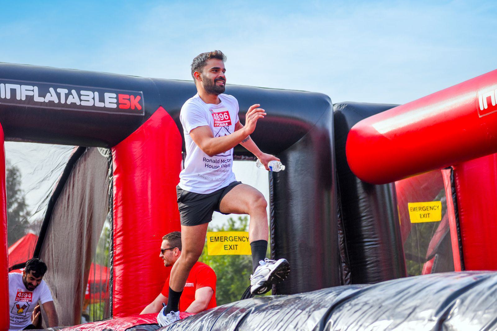 A person wearing a white shirt and black shorts runs on an inflatable obstacle course. Bright red and black inflatables surround them, with visible emergency exit signs. The sky is clear and blue.