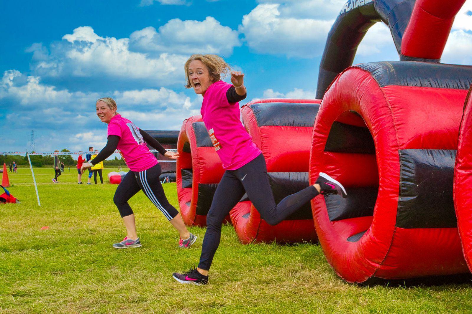 Two women in pink shirts and black leggings are running through an inflatable obstacle course on grass. They look excited and are mid-stride with one smiling and raising her hand. The sky is blue with scattered clouds.