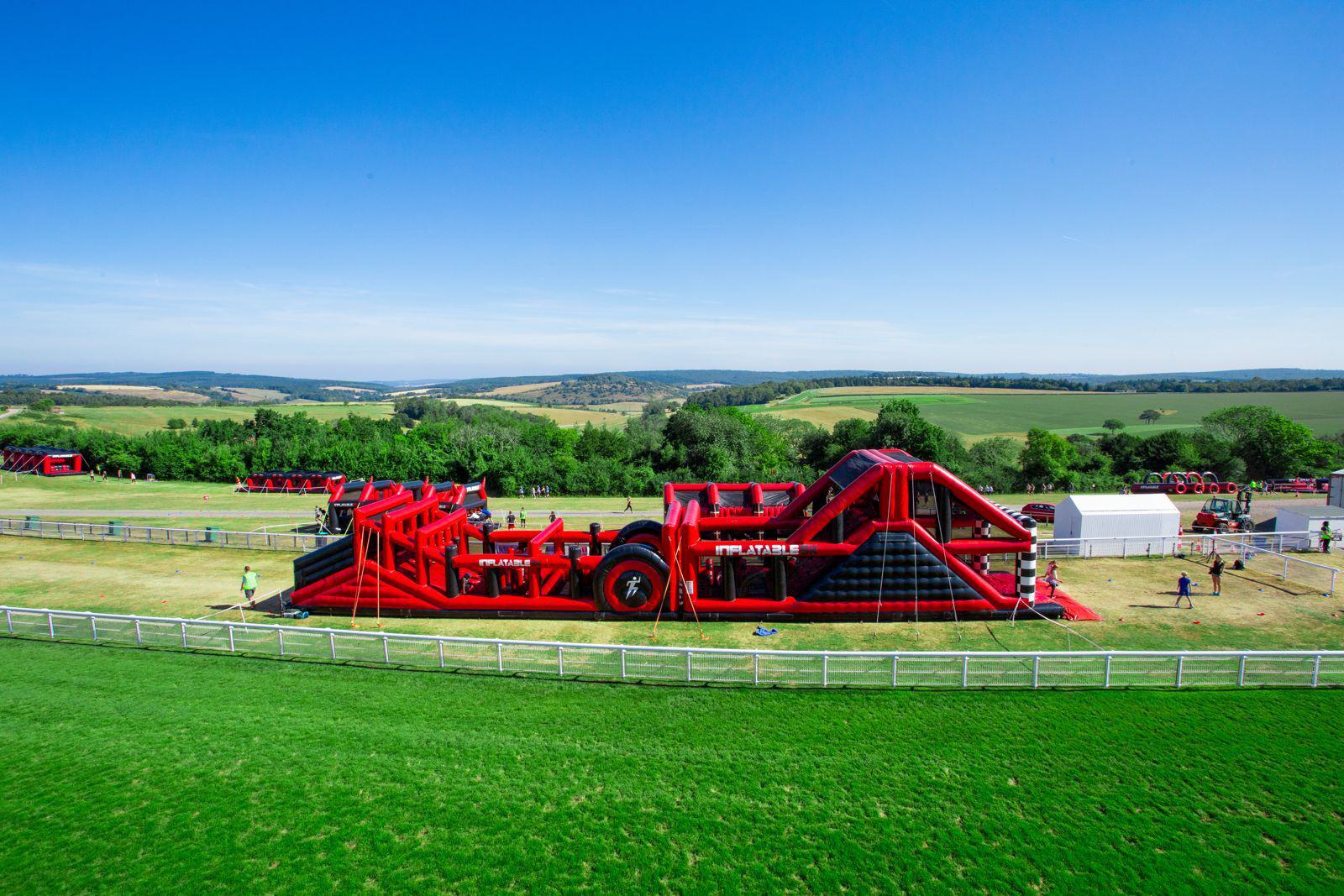 A large red inflatable obstacle course is set up on a grassy field. The course has slides, climbing walls, and tunnels. People are seen engaging with the obstacle course. Rolling hills and green fields form the background under a clear blue sky.