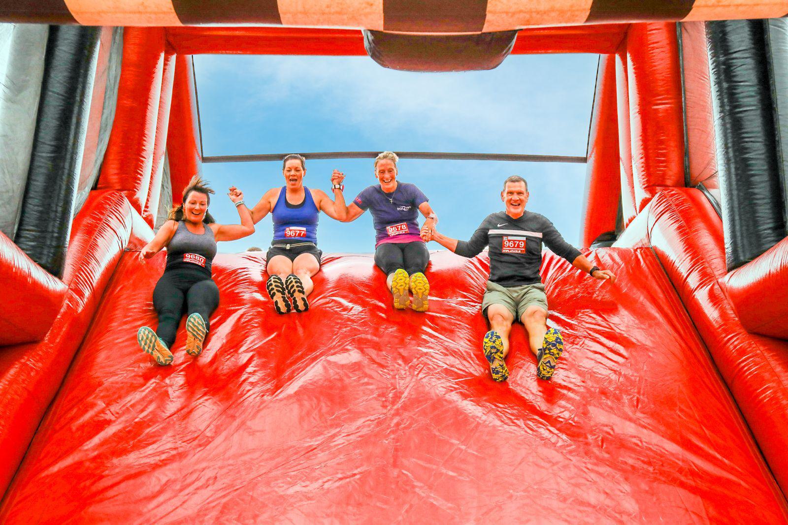 Four people holding hands slide down a large, red inflatable slide. They are smiling and wearing athletic clothes with numbered bibs. The sky is clear and blue above them.