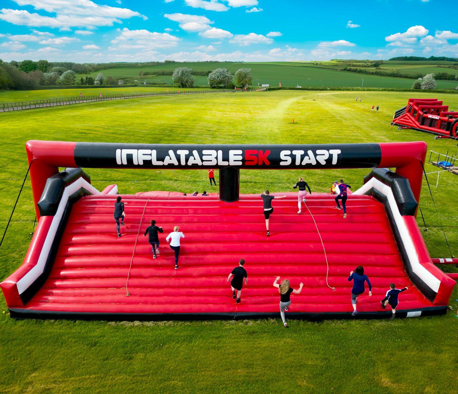Participants race on a giant red inflatable obstacle course under an arch labeled "Inflatable 5K Start." The course is set on a grassy field with expansive green landscape and a blue sky in the background.