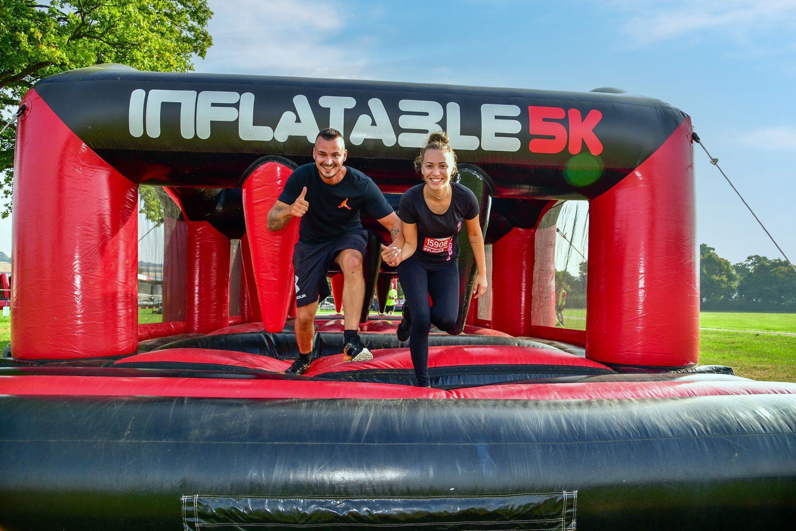 Two people smiling and running on a large inflatable obstacle featuring a red and black design with "Inflatable 5K" written above them. They appear to be enjoying an outdoor fun run event on a sunny day.