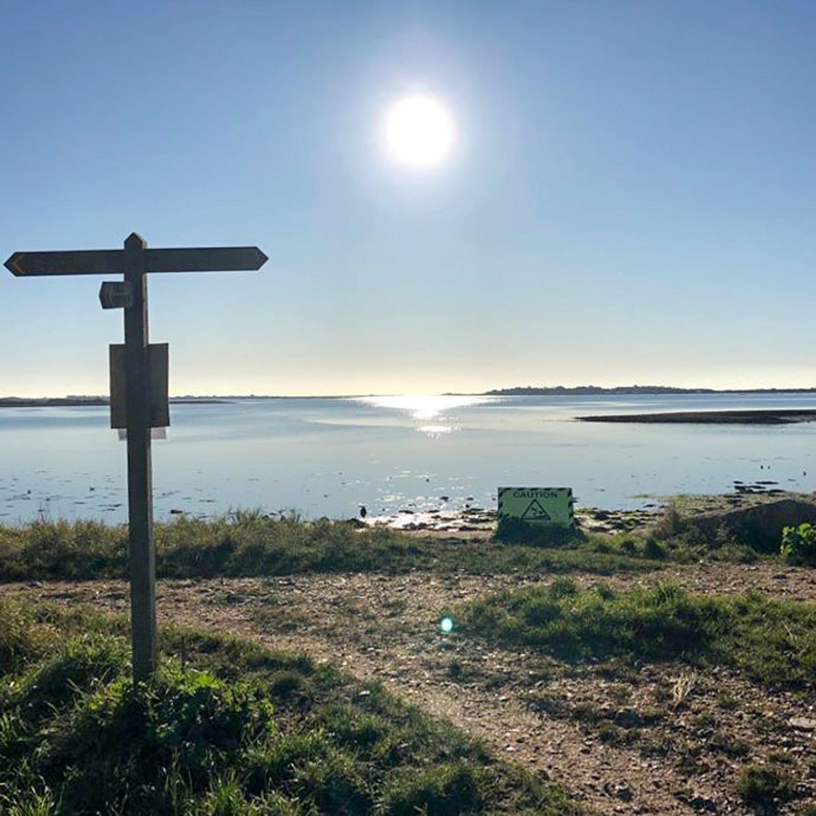 A signpost stands near a serene lakeside path under a clear blue sky with the sun shining brightly. The lake reflects the sunlight, and a small caution sign is visible on the grassy shore.