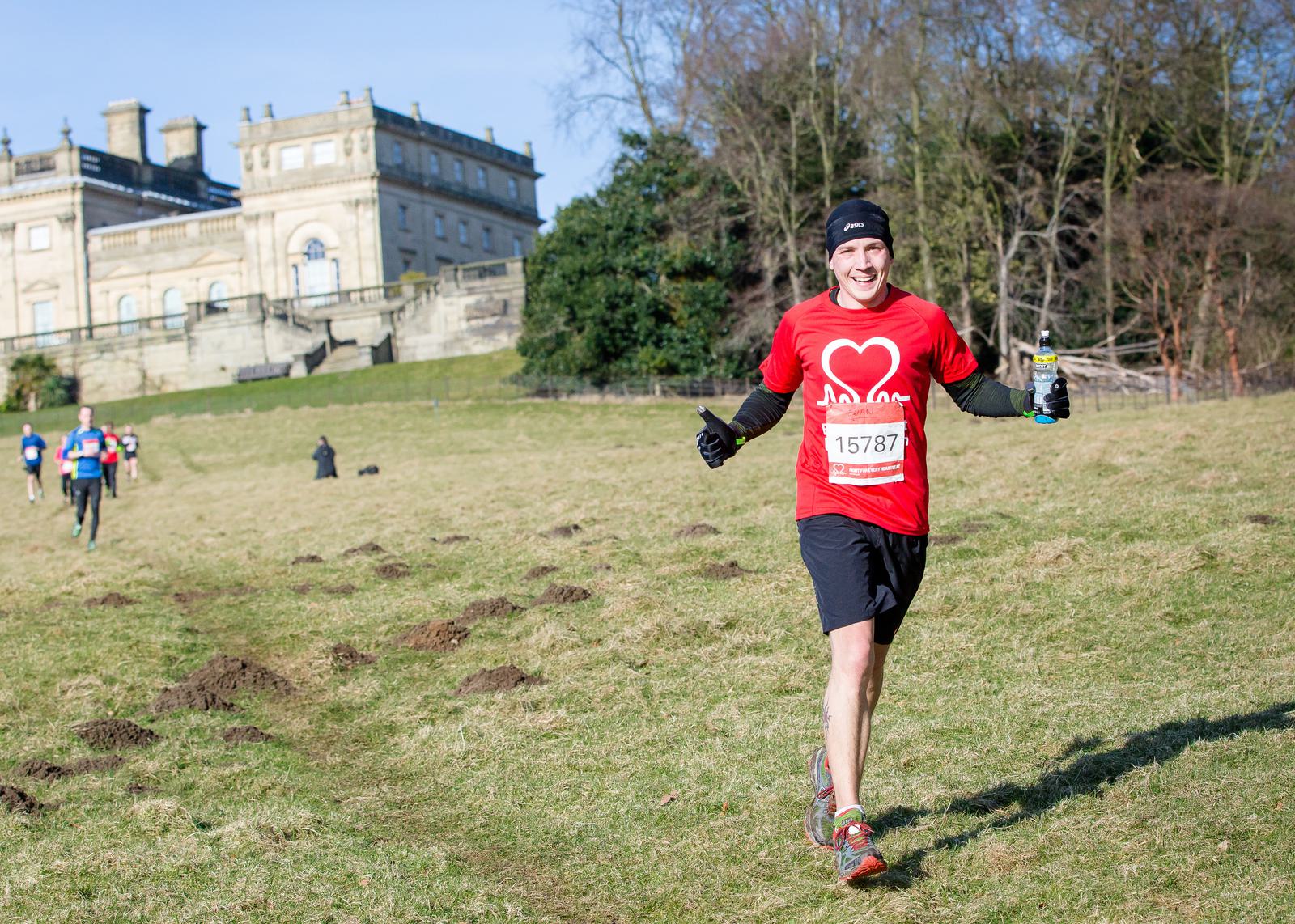 A runner wearing a red shirt, black shorts, and black gloves jogs toward the camera with a smile, holding a bottle in one hand. He's in a grassy field with a historic building and other runners in the background. The runner's race number is 15787.