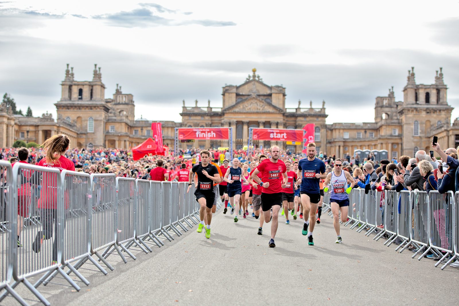 Runners in a race are passing the starting line in front of a large historic building with ornate architecture. Spectators stand on either side of the course, cheering. The sky is overcast, and there are red banners indicating the start and finish lines.