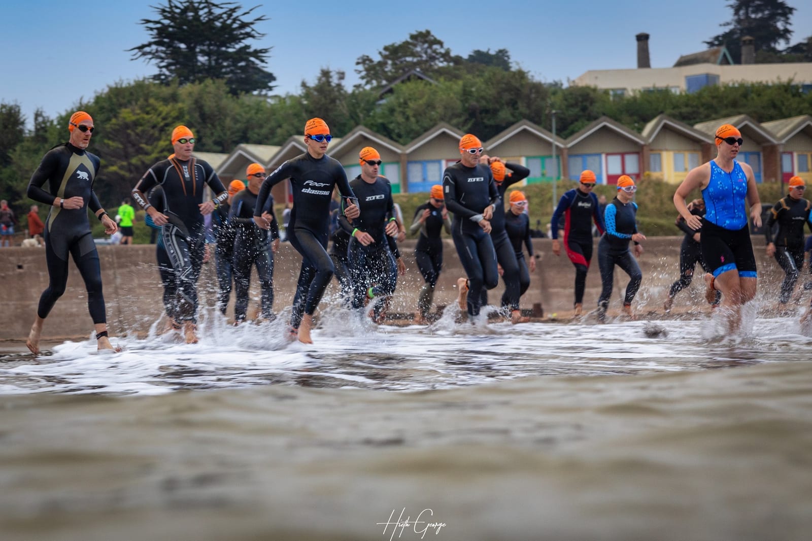 A group of athletes wearing black wetsuits and orange swim caps run into the ocean from the shore. Beach huts and spectators are visible in the background. Some athletes are splashing through the shallow water as they begin their swim portion of a race or triathlon.