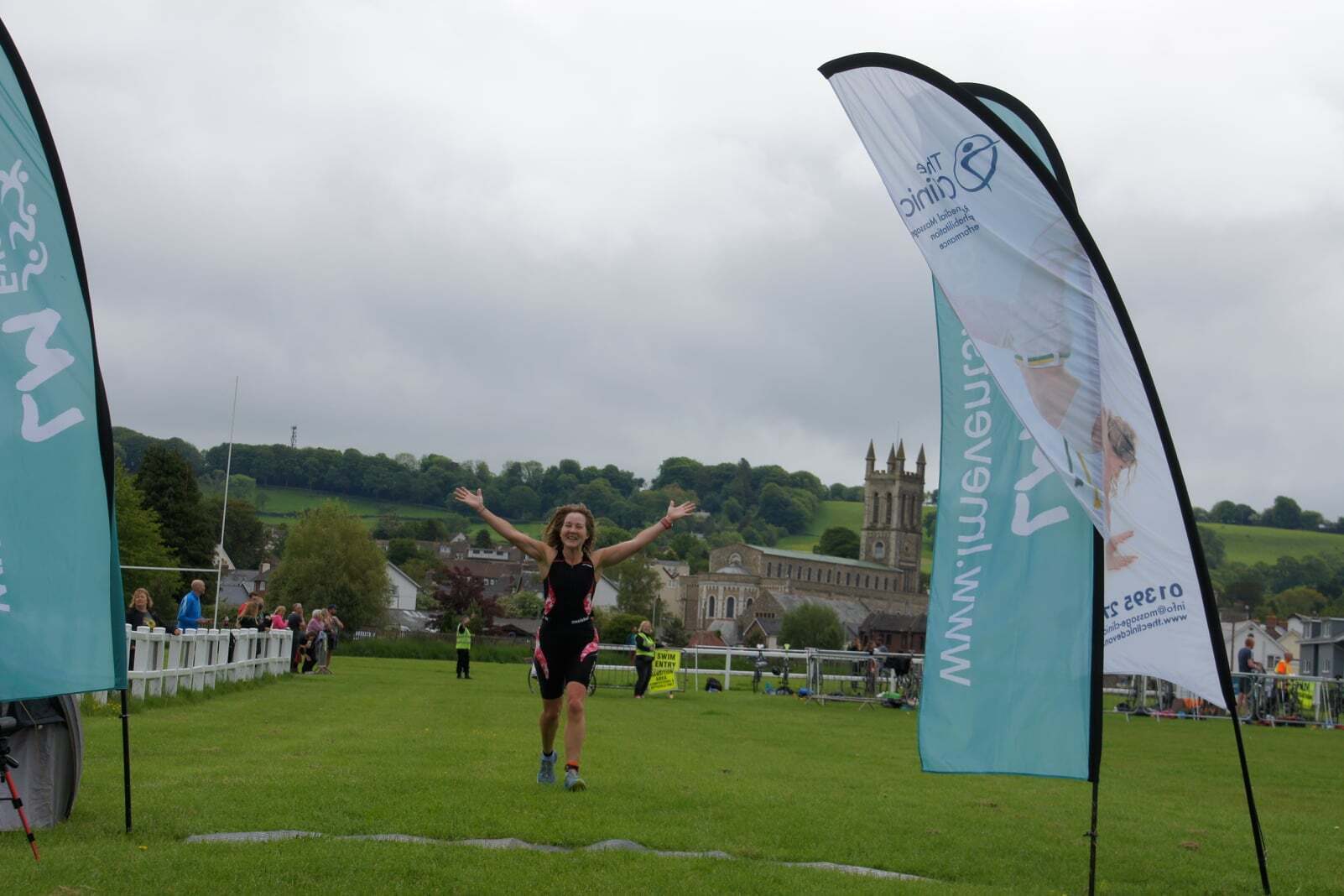 A person wearing athletic gear crosses a grassy finish line with arms raised in celebration. Two flags with advertising text are on either side of the finish line. A historic church stands in the background against a cloudy sky with a wooded hill behind it.