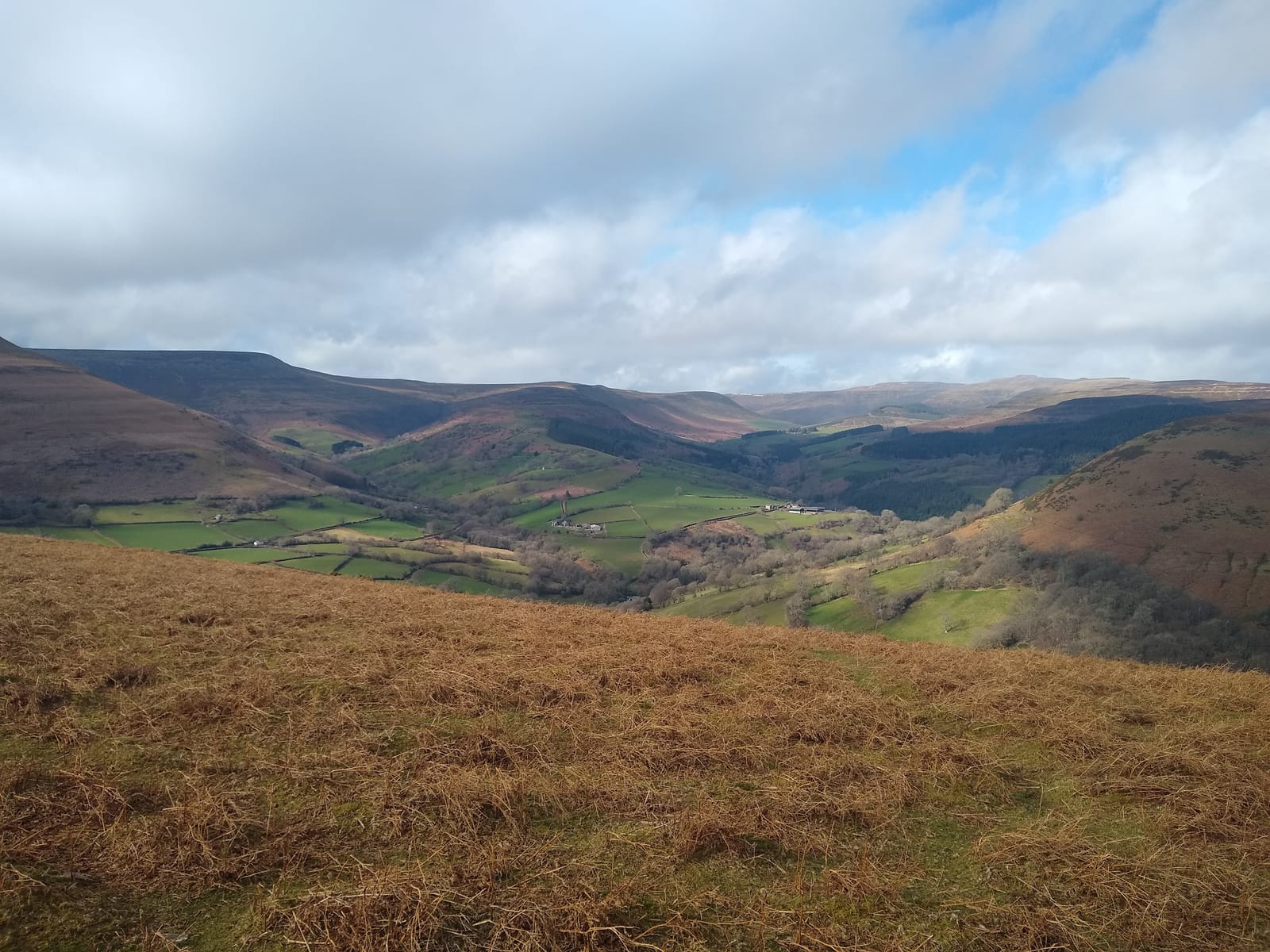 A panoramic view of a serene landscape featuring rolling hills covered in patches of green grass and brown shrubbery. The sky is partly cloudy with some blue visible, and a valley can be seen in the distance with scattered trees and fields.