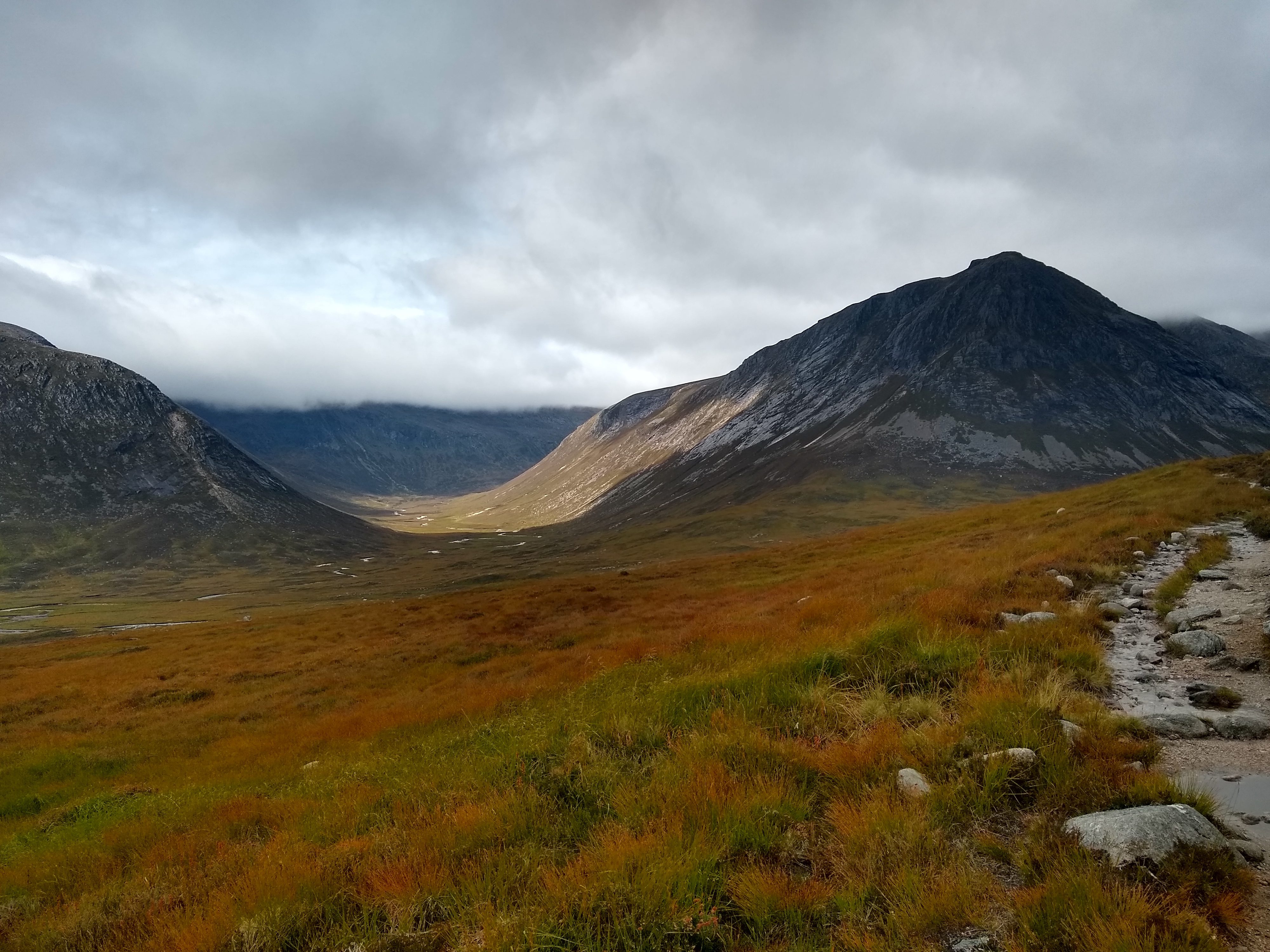 A scenic view of a mountainous landscape with a mix of orange, yellow, and green vegetation. A path winds through the foreground on the right, leading towards tall mountains partially veiled by clouds in the distance. The sky is overcast.