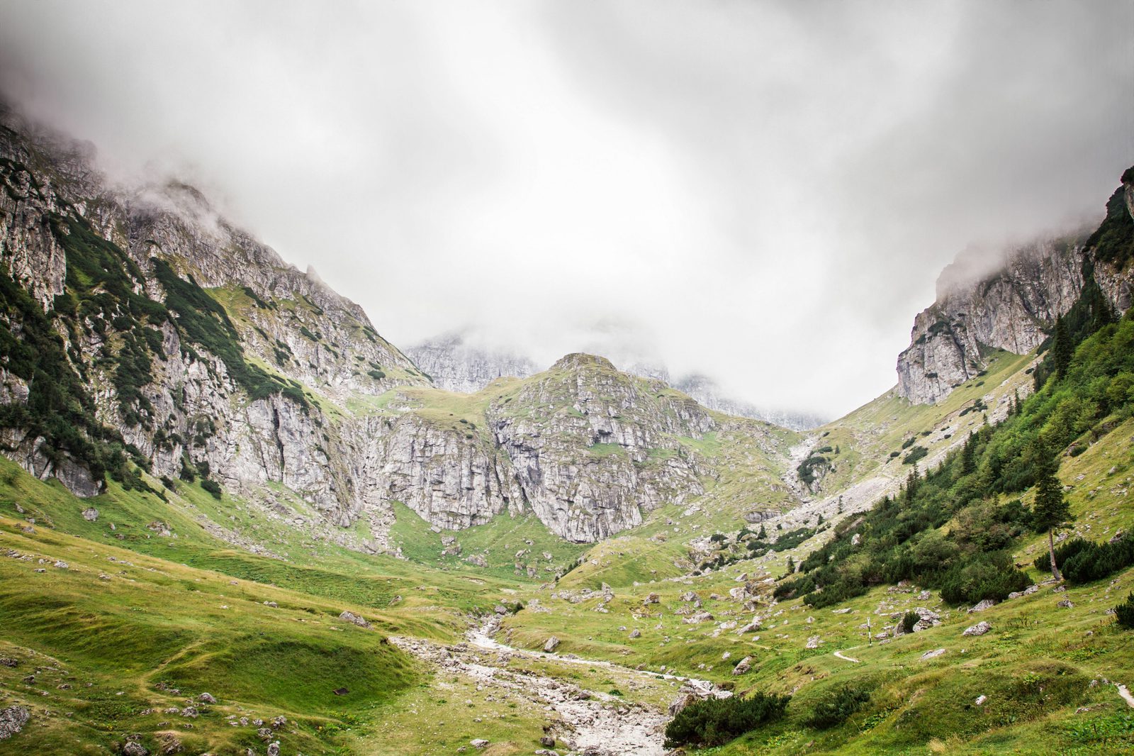 A scenic valley with rocky cliffs and green slopes under a cloudy sky. The landscape includes scattered rocks and patches of grass, presenting a rugged yet serene natural view. The high cliffs are partly shrouded in mist, adding to the tranquil atmosphere.