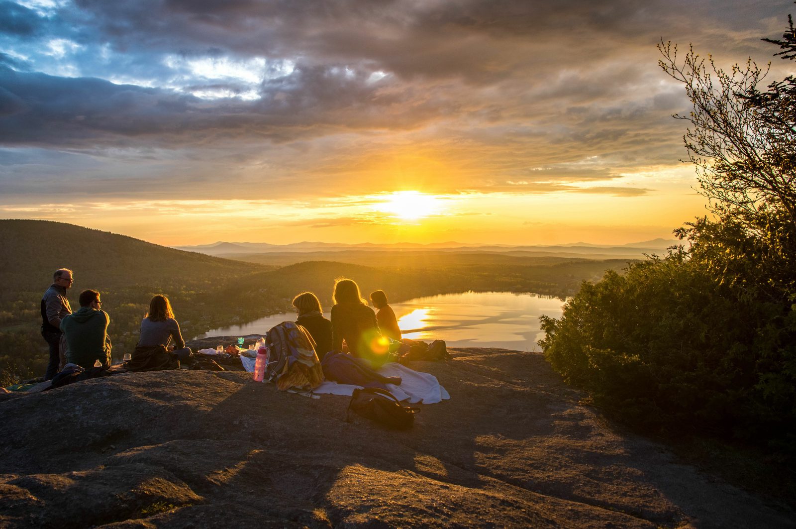 A group of people sitting on a rocky hilltop, watching the sunset over a scenic landscape with hills and a lake. The sky is filled with clouds and the sun creates a golden reflection on the water. The group has blankets and picnic items spread out beside them.