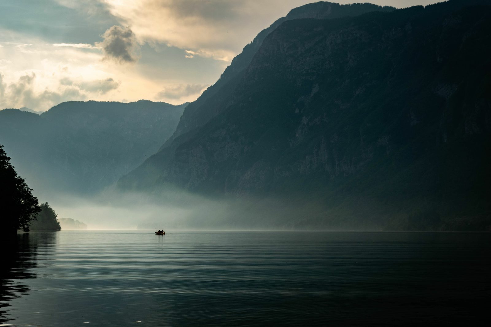 A serene landscape of a calm lake surrounded by towering mountains. Mist rises from the water, creating a mystical ambiance. A small boat with two people is in the distance, silhouetted against the backdrop of the misty mountains. The sky is partly cloudy.