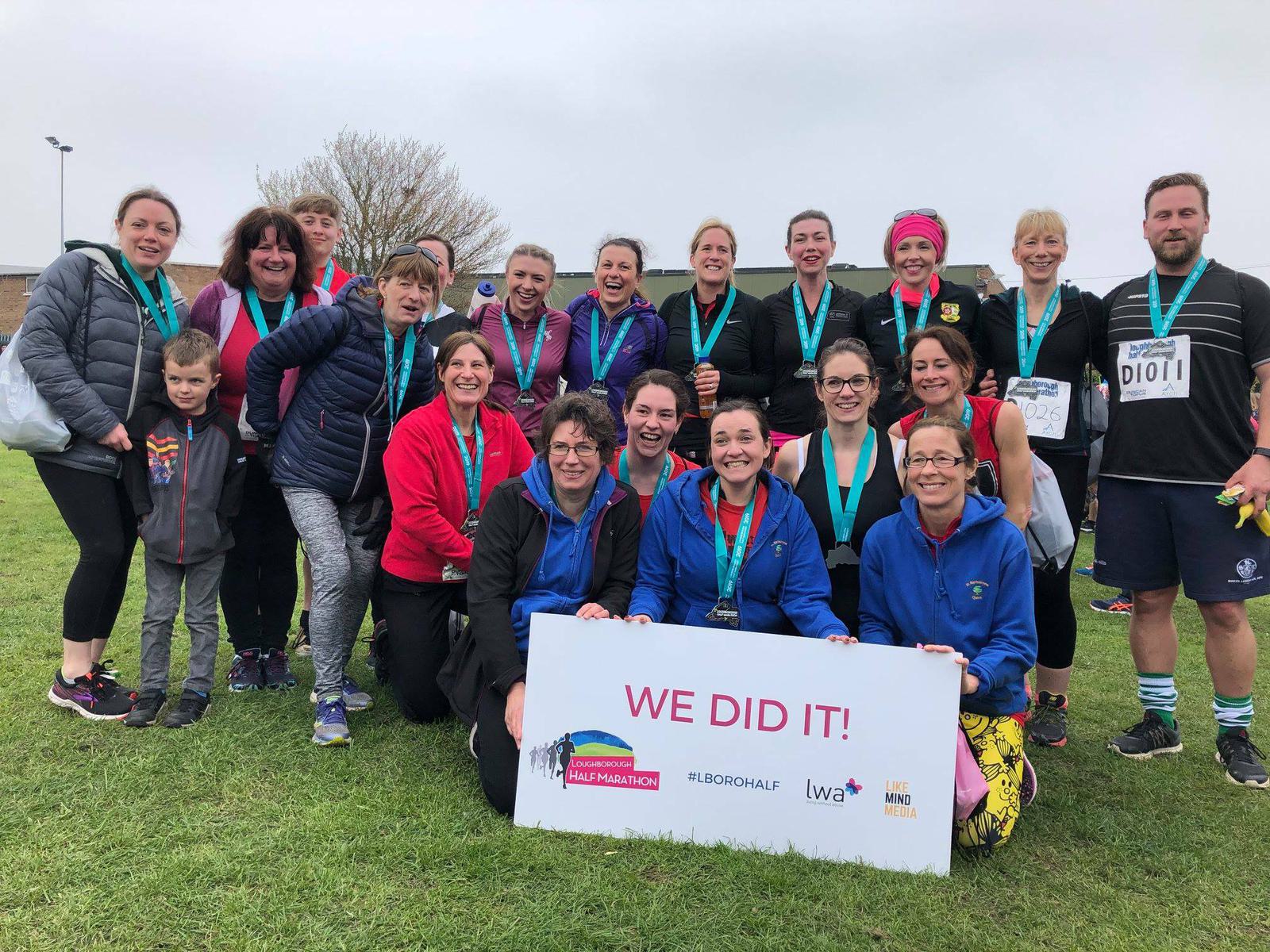 A group of smiling individuals pose outdoors with medals around their necks. They are holding a large sign that reads, "We Did It!" and includes several logos. The individuals appear to be celebrating the completion of an event, likely a race.