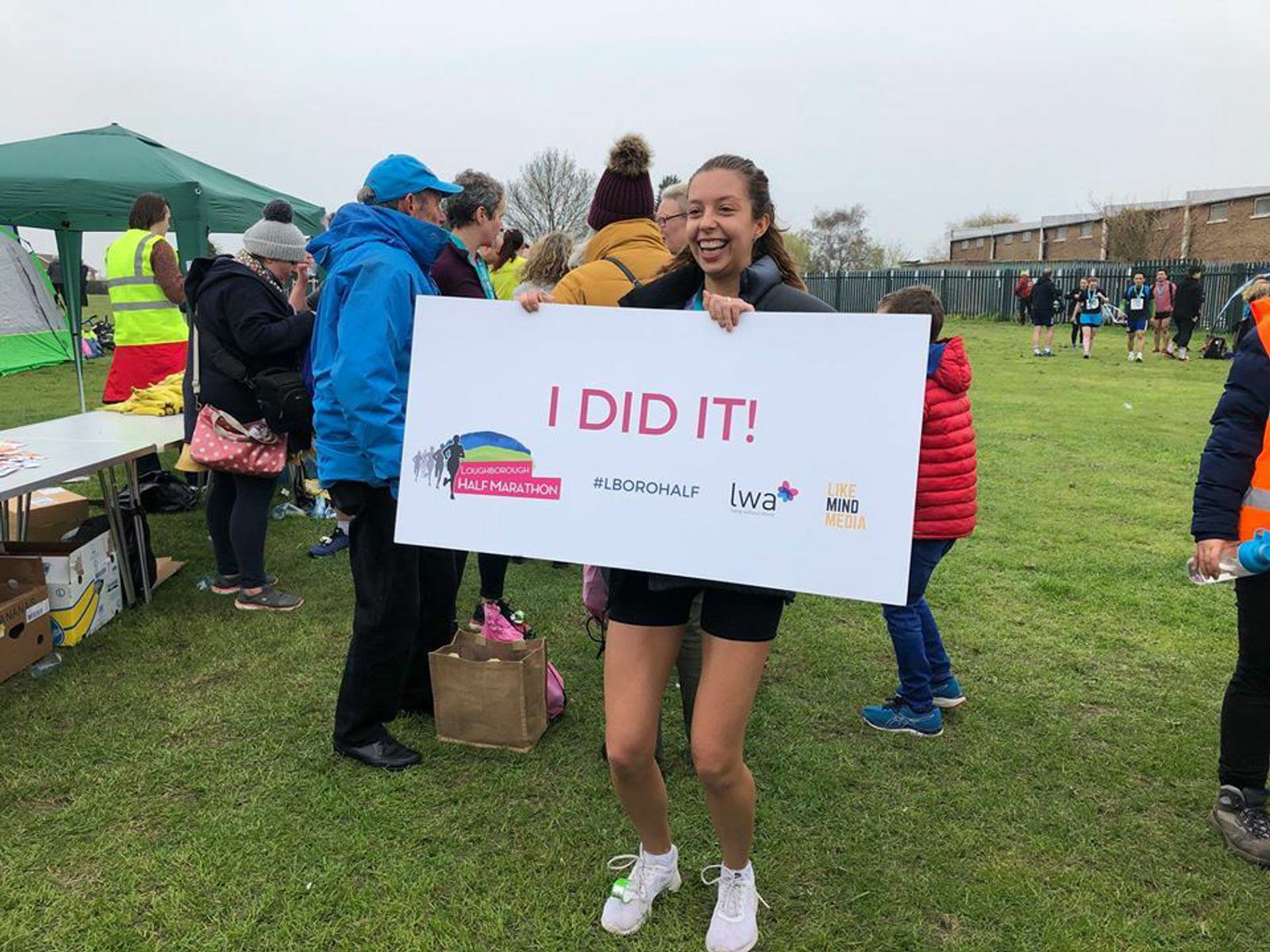 A woman stands on a grassy field, smiling broadly while holding a large sign that reads "I DID IT!" She is wearing athletic clothing and white sneakers. People and a few tents can be seen in the background, indicating a race or marathon event.