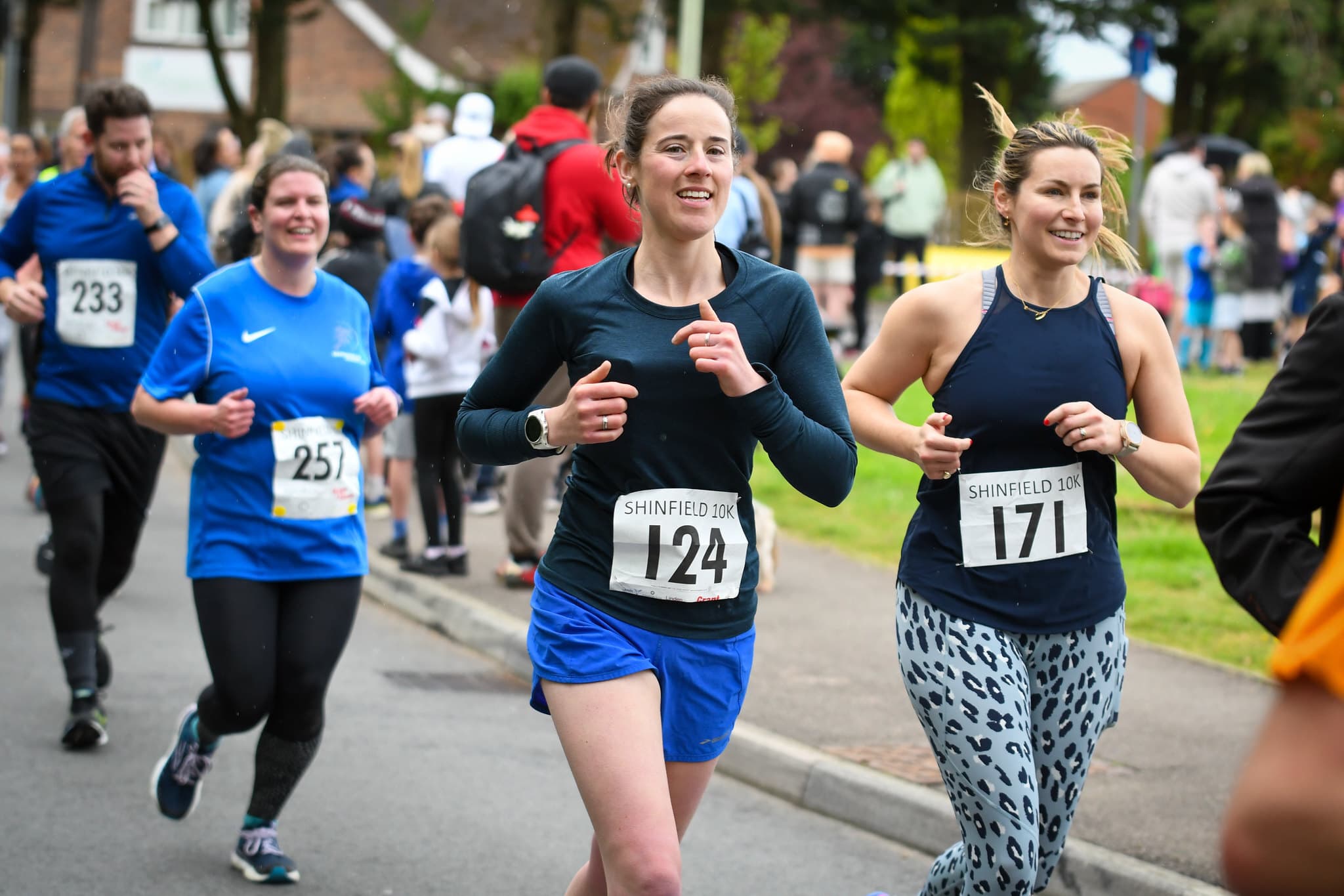 Runners participating in a road race, smiling and wearing numbered bibs. The focus is on a woman in a navy shirt and blue shorts, bib number 124, running alongside others on a street lined with spectators.
