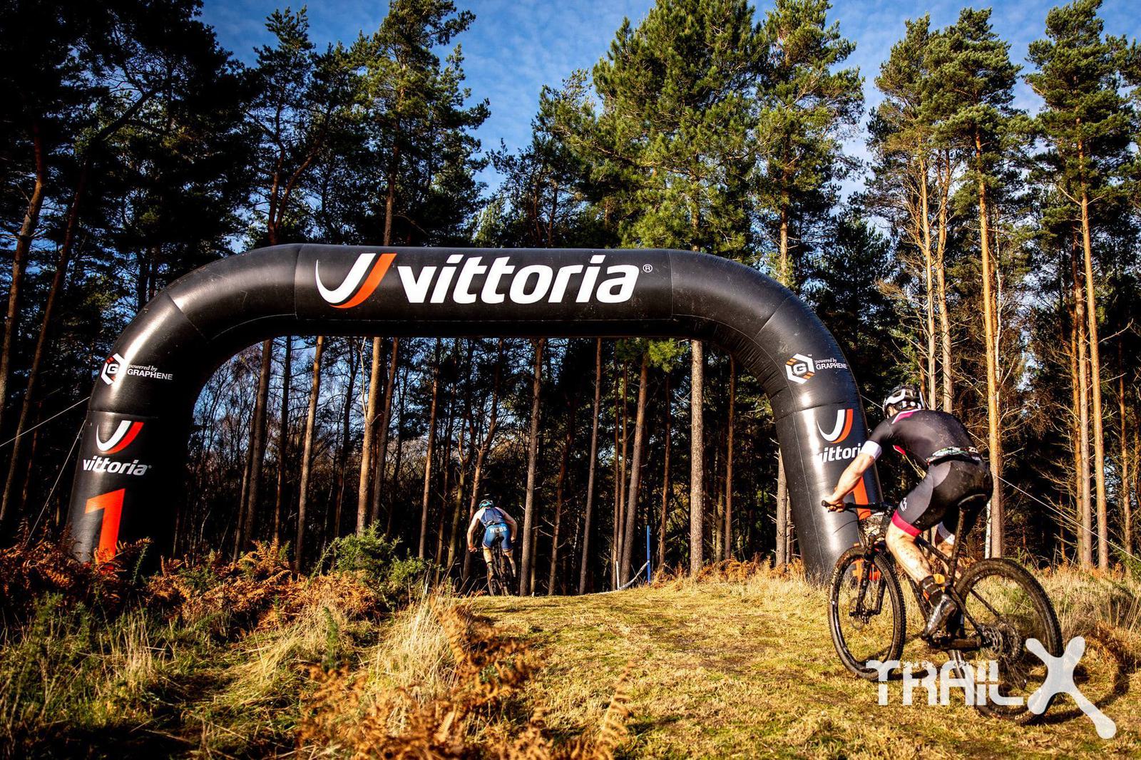 Mountain bikers riding under a black inflatable arch with the "Vittoria" logo on a forest trail. The terrain is grassy with some ferns, and the area is surrounded by tall trees under a clear blue sky. The "TrailX" logo is visible in the lower right corner.
