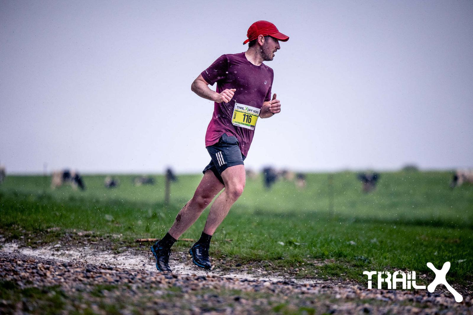 A man running outdoors in the rain on a gravel path. He is wearing a red cap, a dark t-shirt, shorts, and a race bib numbered 116. The background is a grassy field with blurred cows and a gray sky. "TRAILX" is visible at the bottom right of the image.