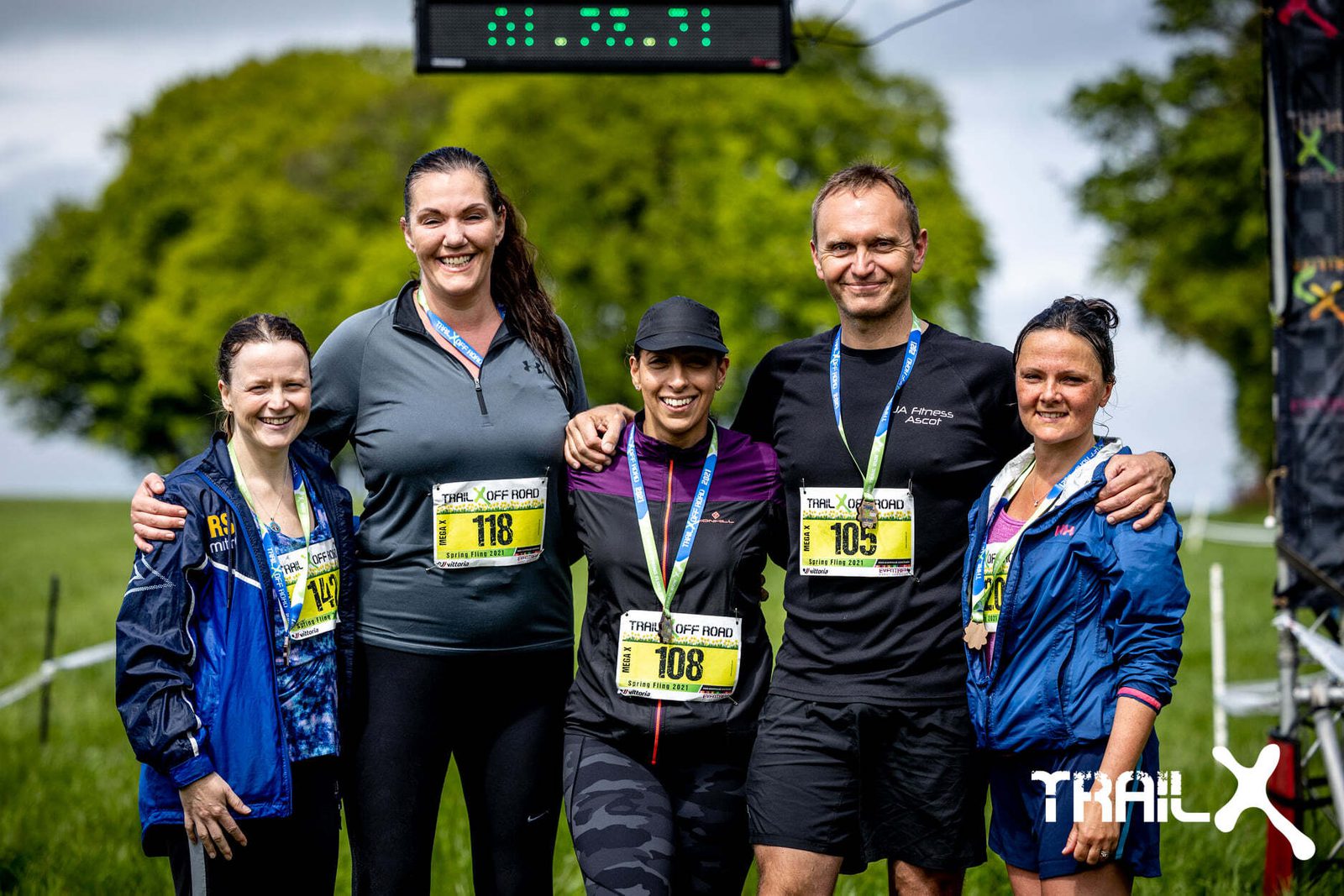 Five smiling runners pose together under a finish line clock with green fields and a large tree in the background. They are wearing race bibs and athletic gear. The logo "TRAILX" is visible in the bottom right corner.