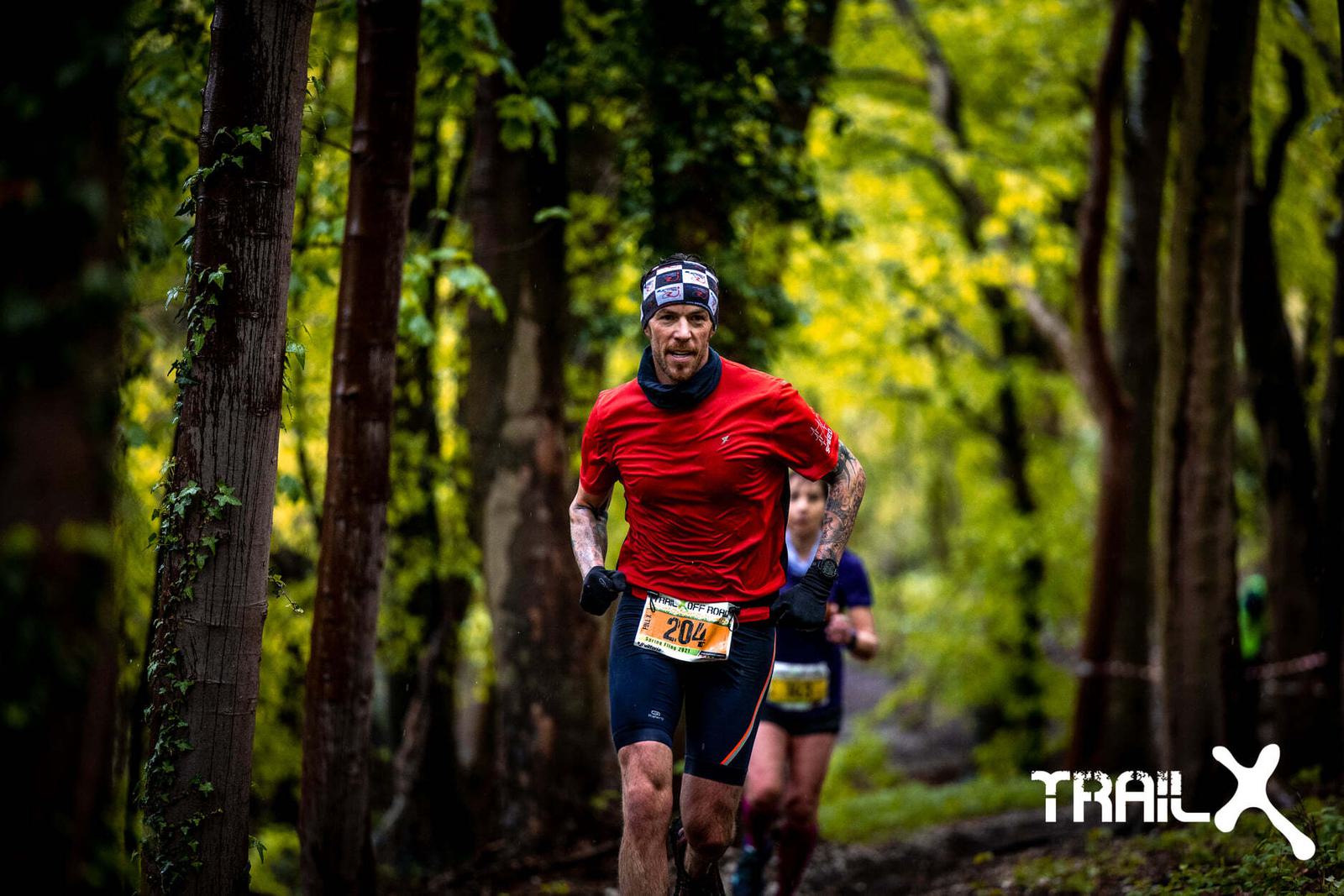 A man wearing a red shirt and a headband is running through a forest trail during a race. He has a race bib number 204 on his shorts. Another runner is visible behind him. The surroundings are lush with green foliage. A logo reading "TRAIL X" is on the bottom right.