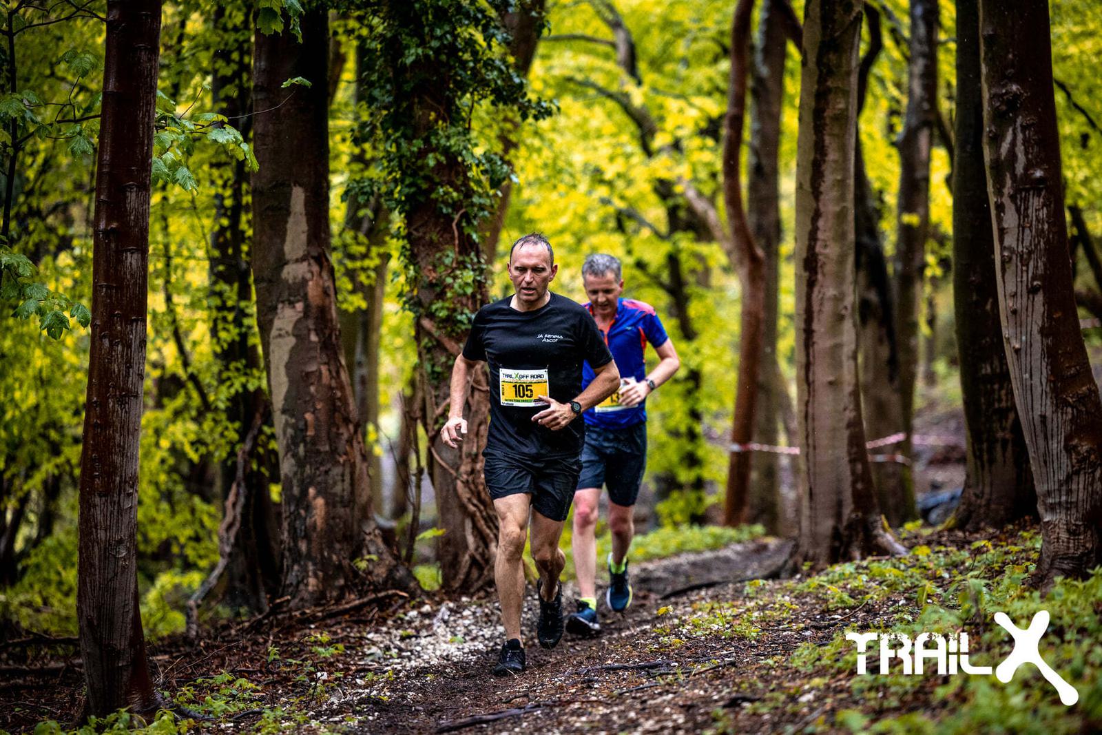 Two men are trail running through a lush, green forest. The runner in front wears a black t-shirt and shorts with the number 116. The other runner is just behind him wearing a blue shirt. The ground is muddy, and the atmosphere appears damp. A logo reading "TRAILX" is in the corner.