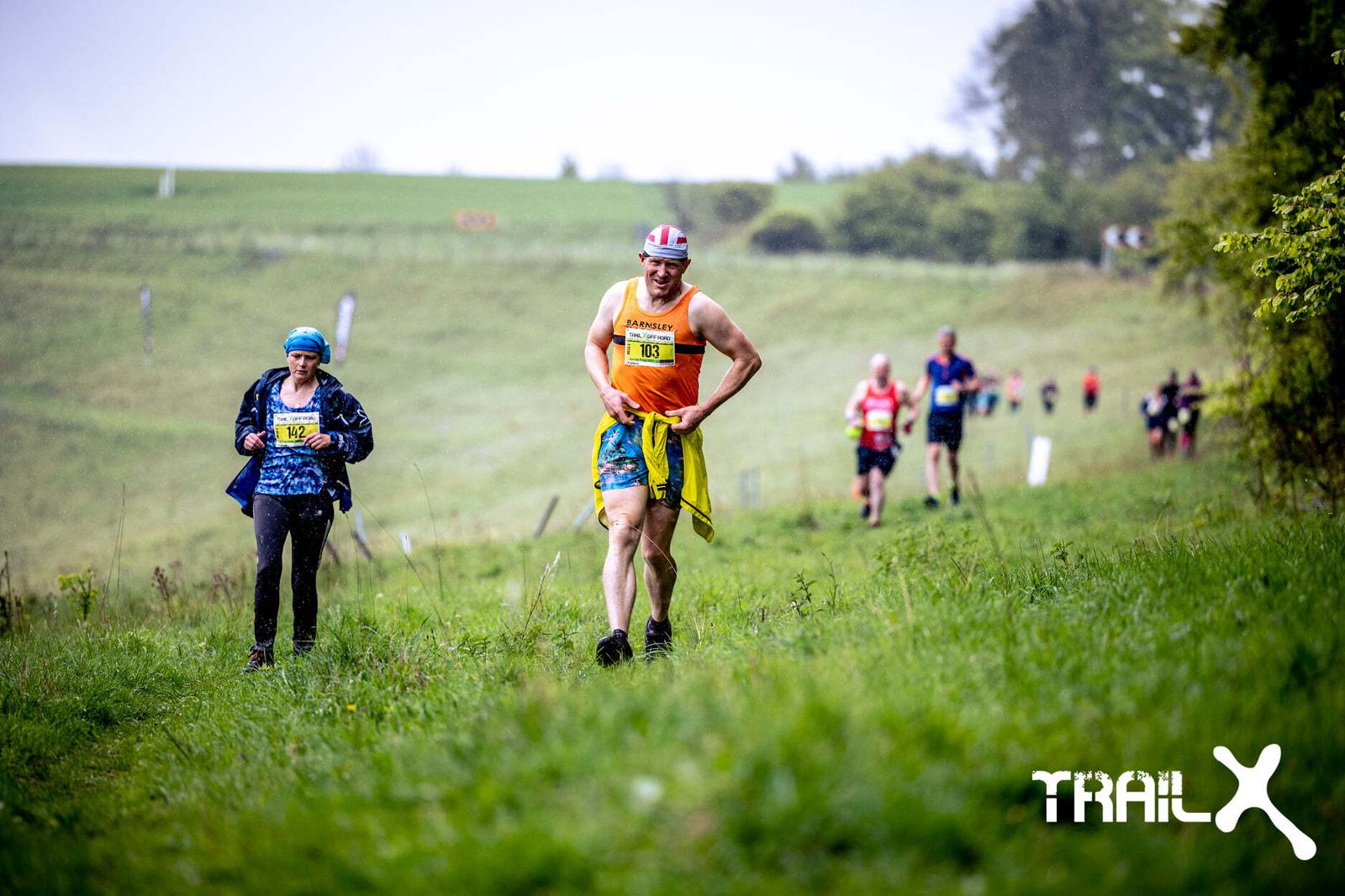 Runners navigate a grassy, slightly uphill trail during an outdoor race, each wearing race bibs. The leading runner wears a colorful outfit and a headband, while the others follow at varying distances. The background is a lush, green countryside with a few scattered trees.