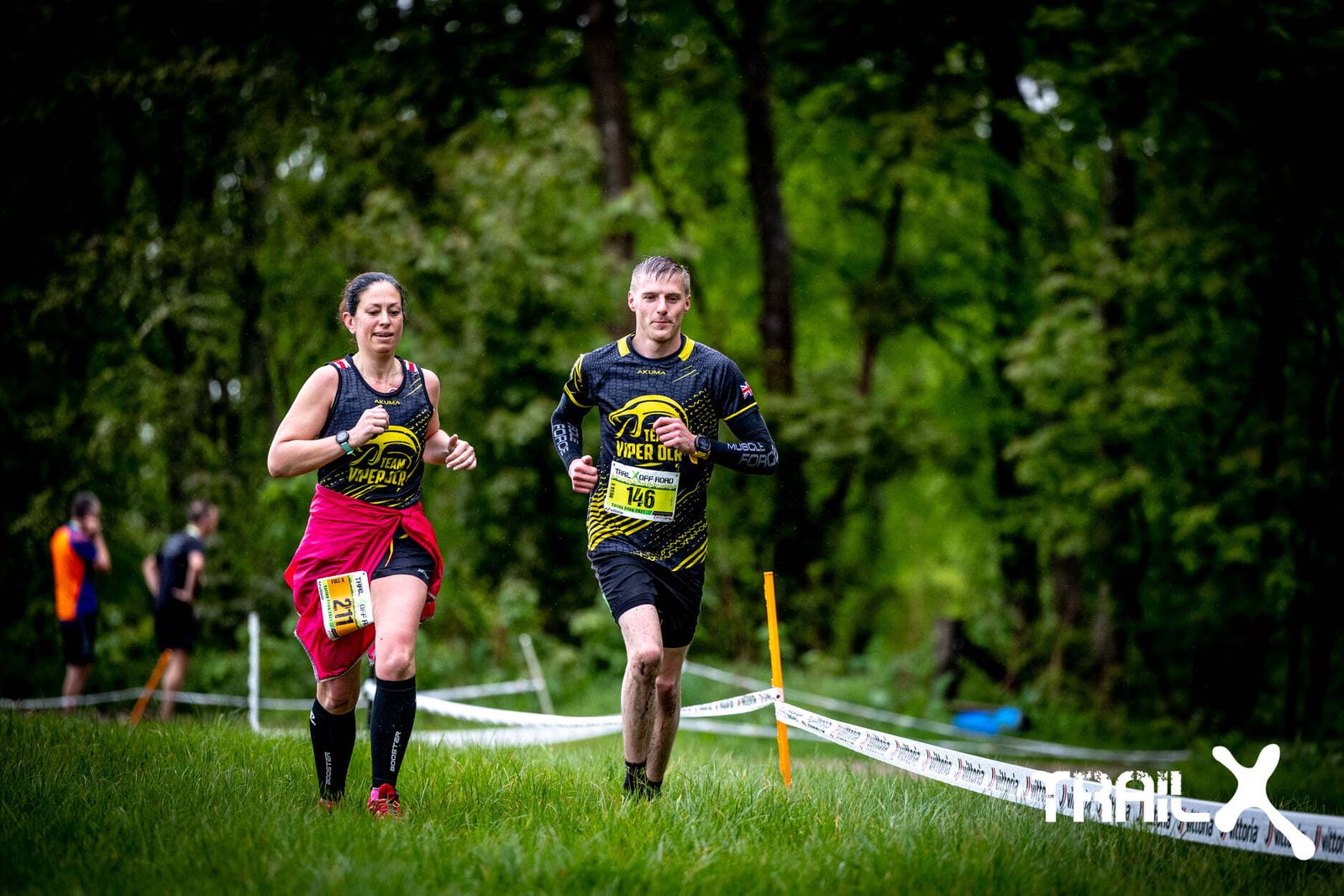 Two runners, a woman and a man, participate in a trail race through a forest. Both wear yellow and black sportswear, with the man donning a race bib number 146. The woman has a pink jacket tied around her waist. Other runners and a white race tape are visible in the background.