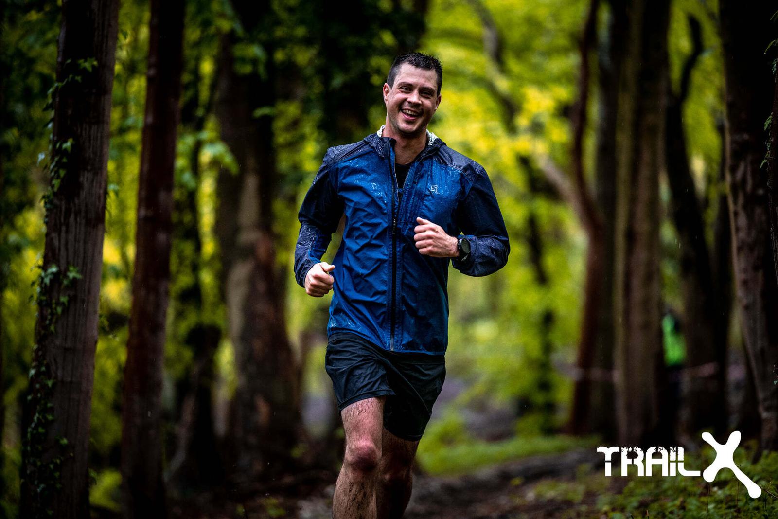 A person, smiling and wearing a blue jacket and black shorts, runs on a forest trail. The surroundings are lush and green, and the ground appears wet, suggesting recent rain. The logo "Trail X" is visible in the lower right corner of the image.